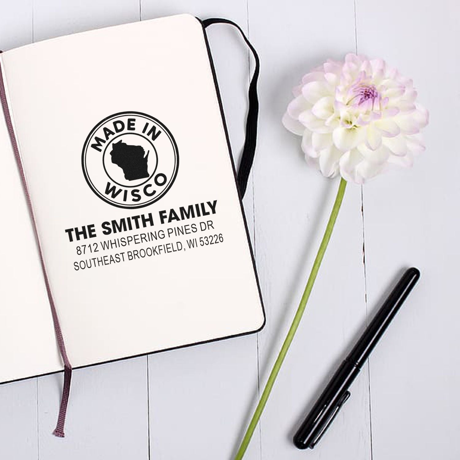 Custom family return address stamp with Made in Wisco design, displayed on an open notebook beside a white flower and black pen on a white wood desk.