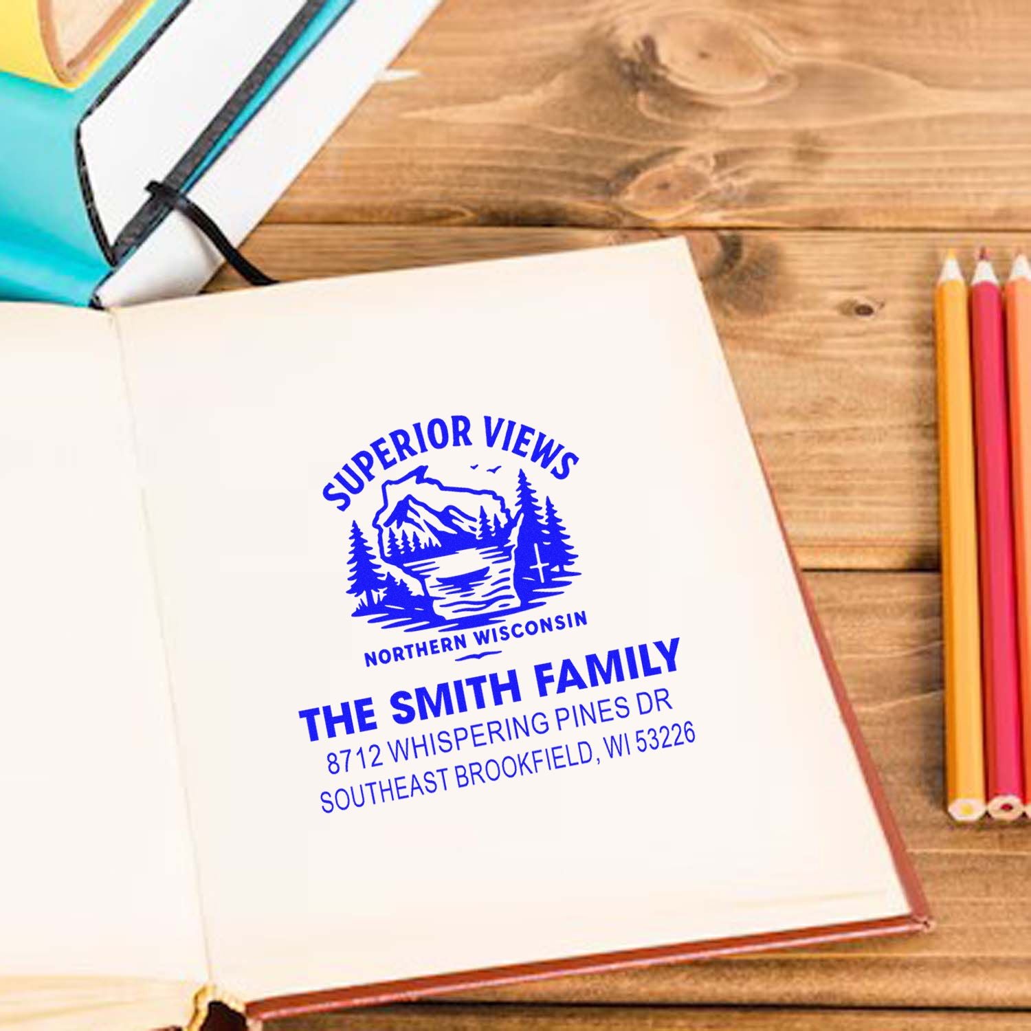 Custom address stamp with Superior Views logo and Smith family address printed on an open notebook, on a wooden desk with pencils and sketchbooks.