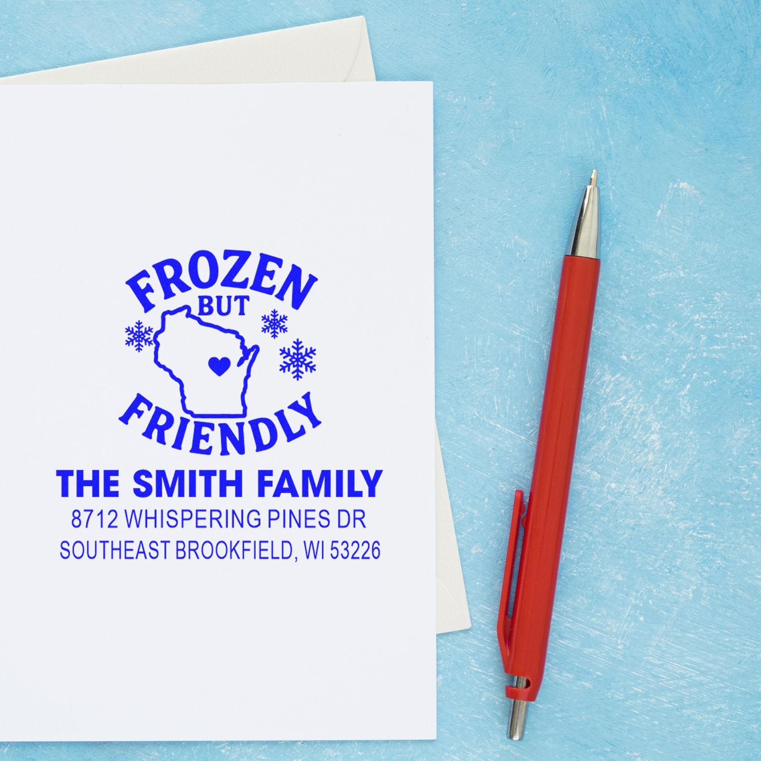 Custom blue return address stamp on white envelope reading Frozen but Friendly, with Wisconsin outline and family address, beside a red pen on blue background.