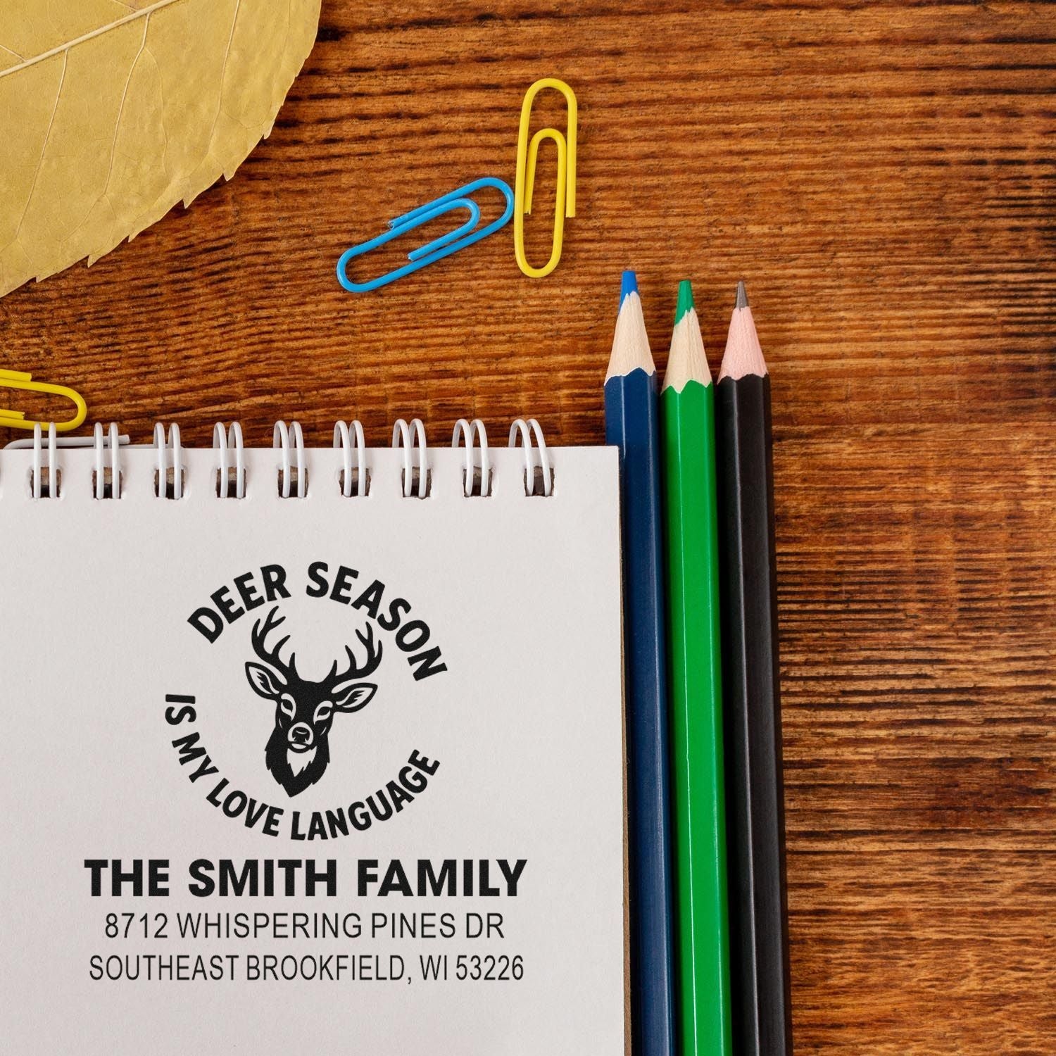 Personalized deer season family address stamp on a spiral notebook, with colored pencils and paper clips on a wooden desk.