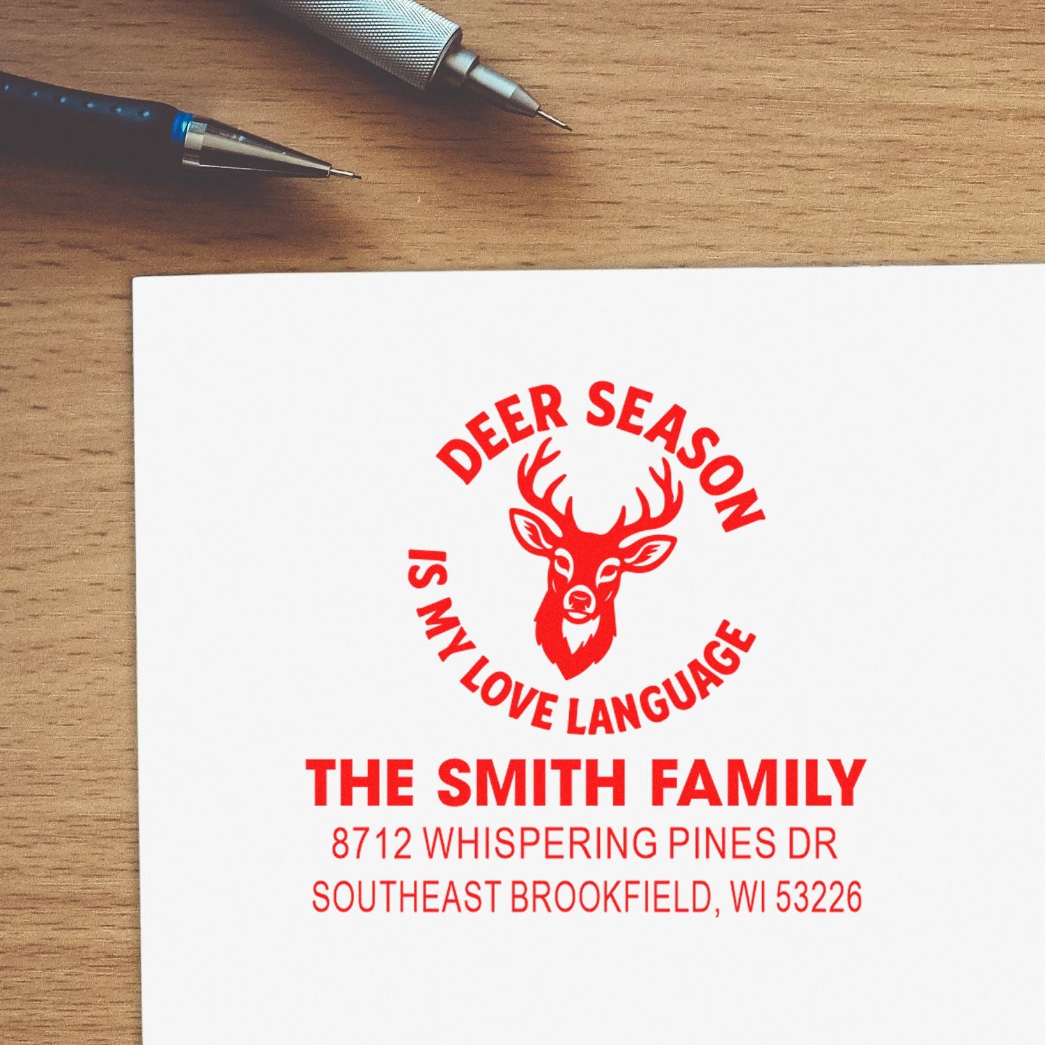 Custom deer season family address stamp on white paper beside pens on a wooden desk, featuring a deer logo and the Smith family address.