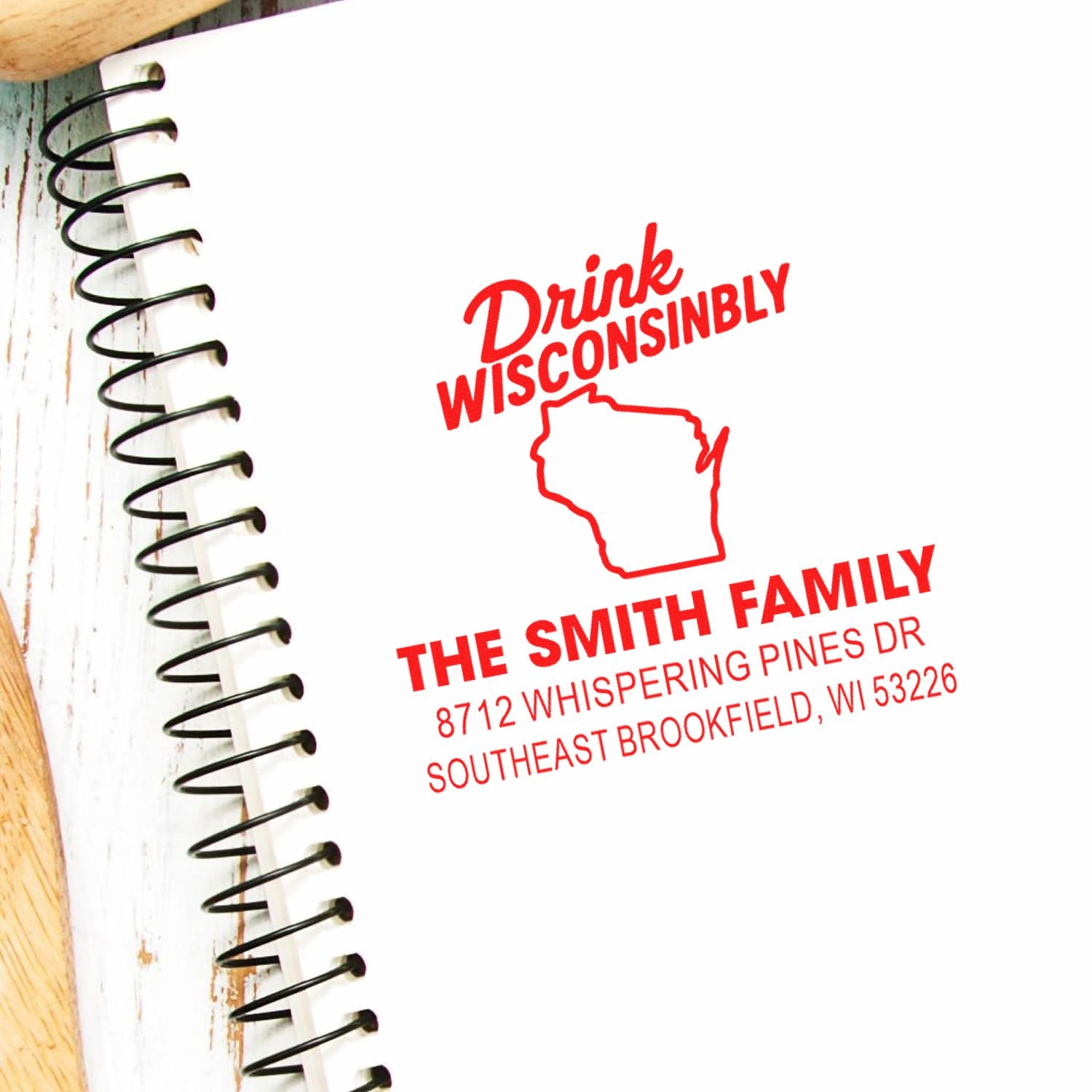 Custom red address stamp on a spiral notebook, reading Drink Wisconsinbly with Wisconsin outline and The Smith Family address.