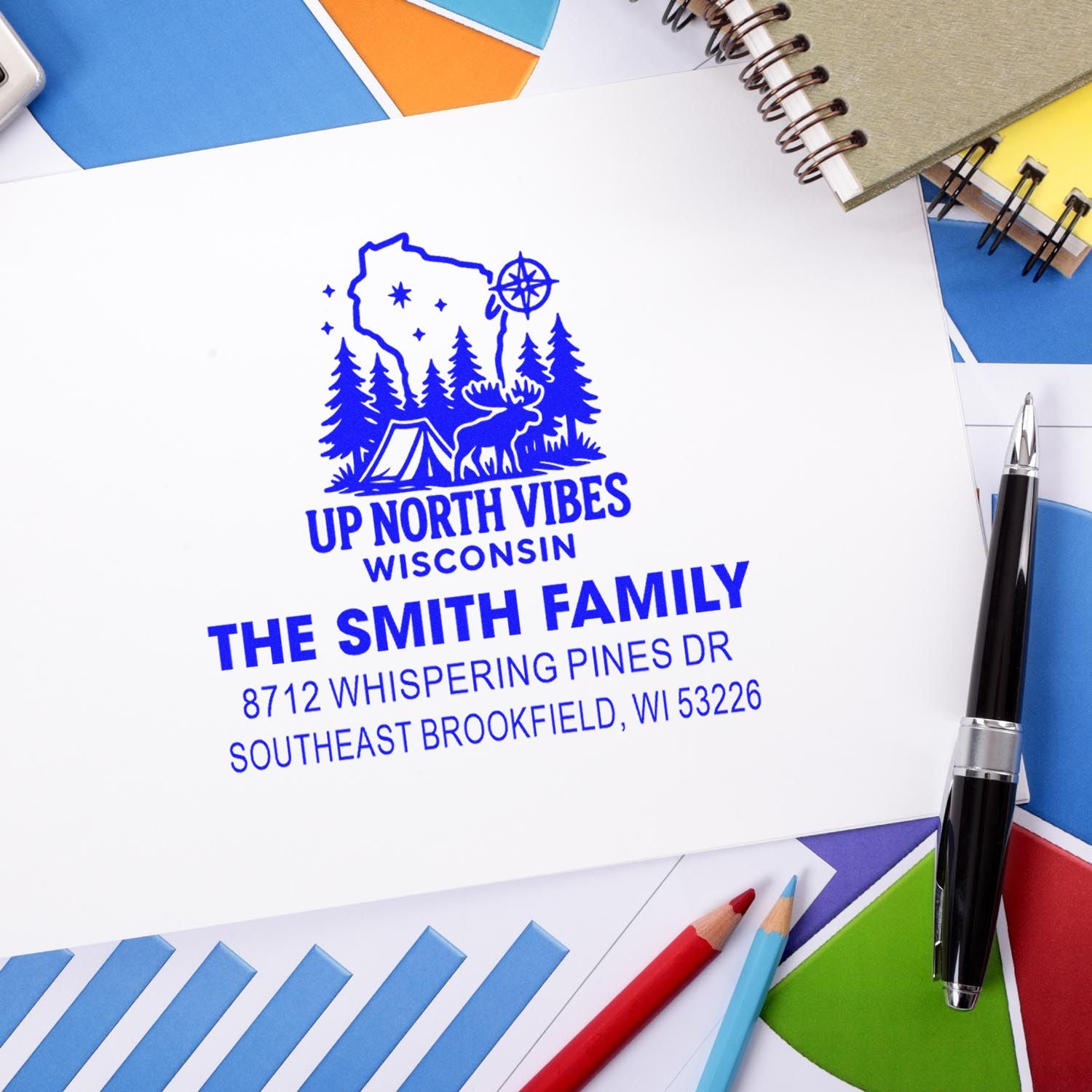 Custom address stamp with Wisconsin camping logo and The Smith Family details on white paper, surrounded by notebooks, pens, and colorful stationery.
