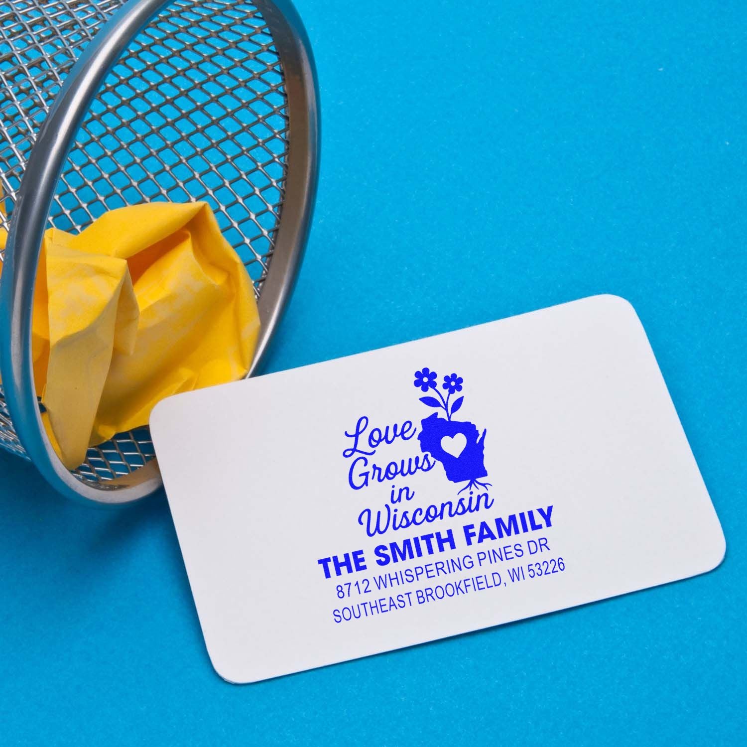 Custom address stamp with Love Grows in Wisconsin design, shown on a blue background beside a metal basket and yellow tissue paper.
