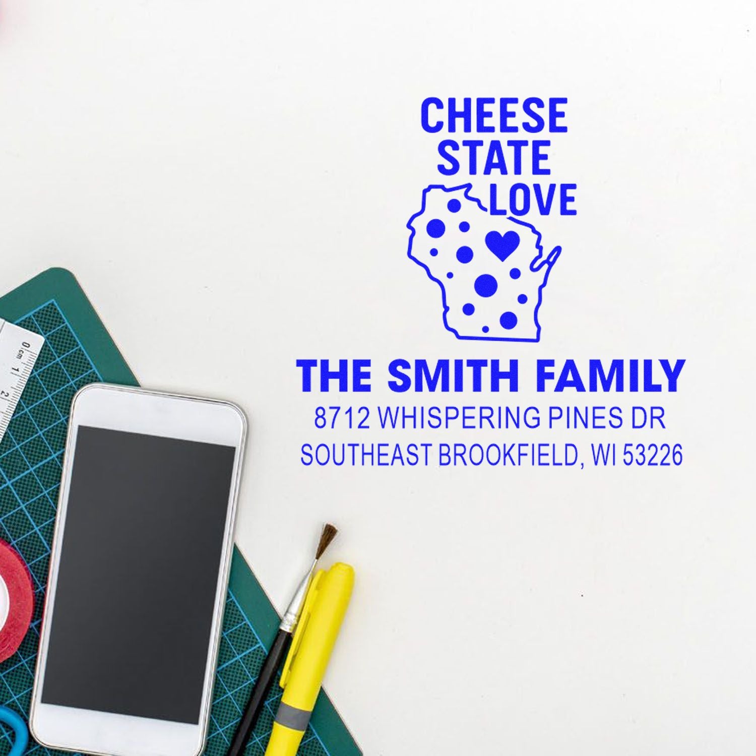 Blue custom return address stamp with Wisconsin outline, Cheese State Love, family name and address on white desk with crafting tools.
