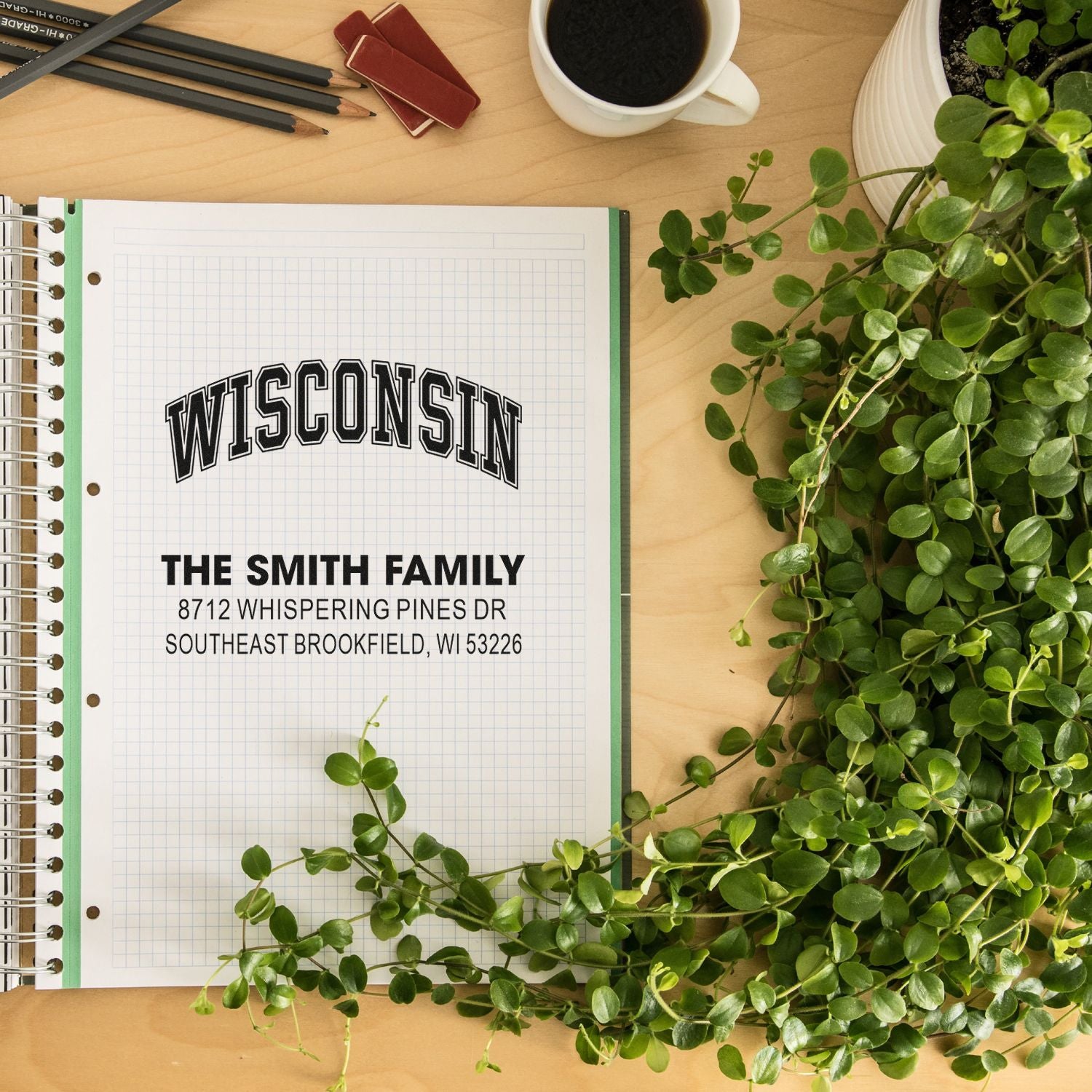 Personalized Wisconsin family address stamp on graph paper notebook, styled on a desk with coffee, pencils, and trailing green plant.