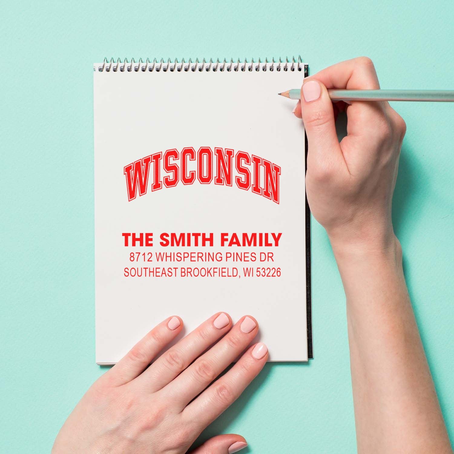 Custom Wisconsin return address stamp on a spiral notebook, held by hands with a pencil on a mint green background.