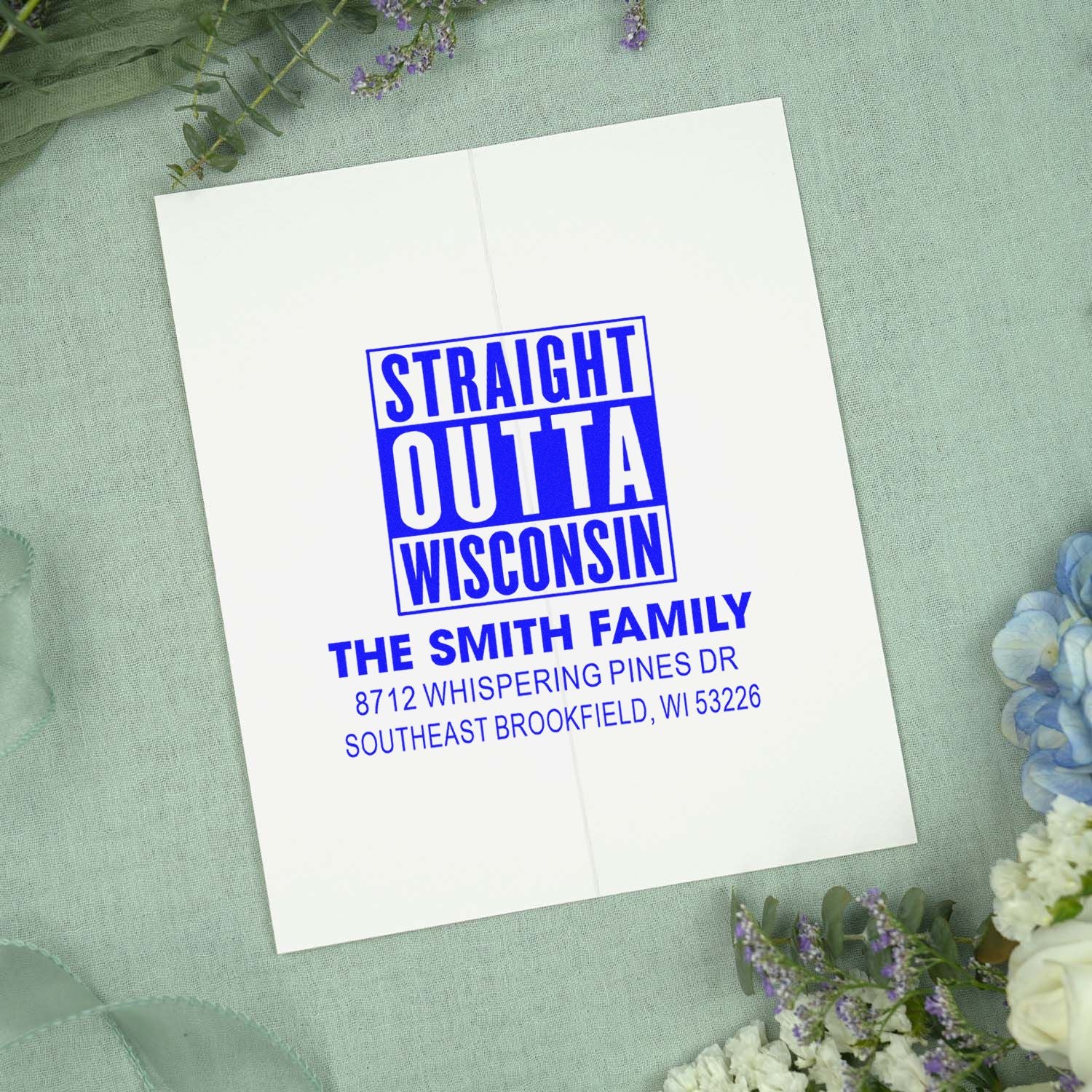 Personalized self-inking address stamp with blue Straight Outta Wisconsin design for The Smith Family on a white card with floral backdrop.