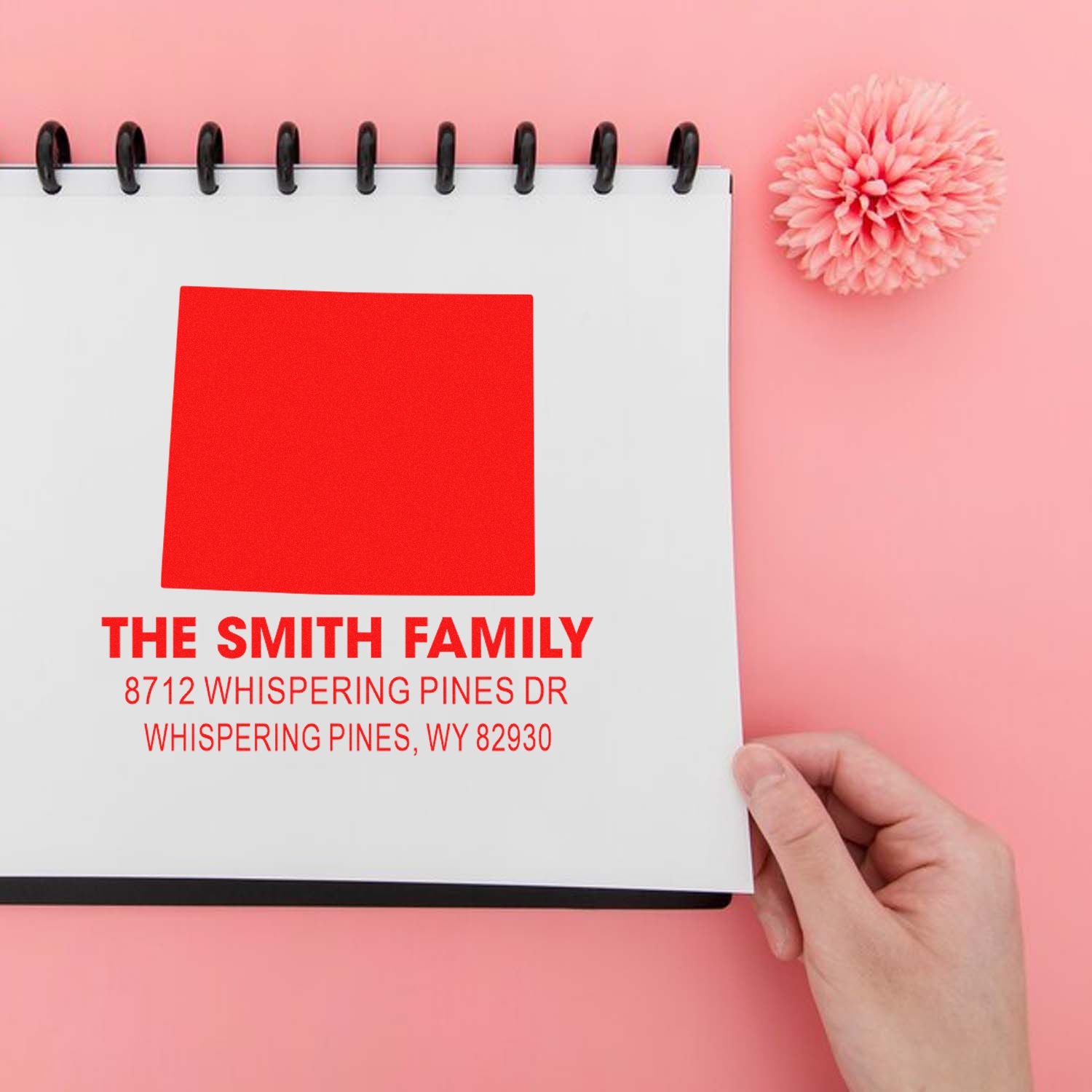 Personalized family address stamp mockup on a spiral notebook with pink background and flower, showing red custom return-address imprint.