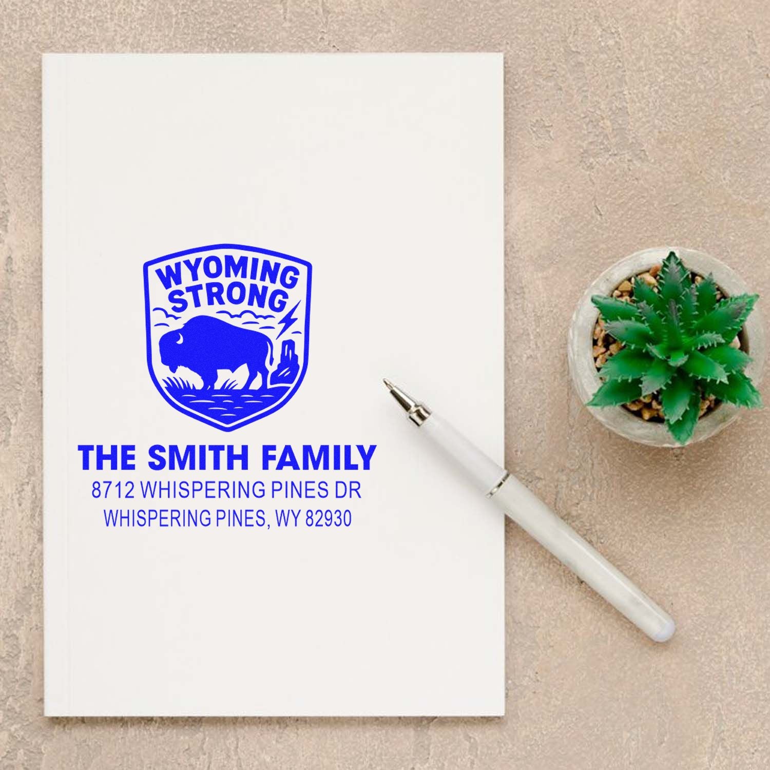 Custom Wyoming Strong self-inking address stamp with bison logo, Smith family name, and Whispering Pines, WY address beside a white pen and succulent.