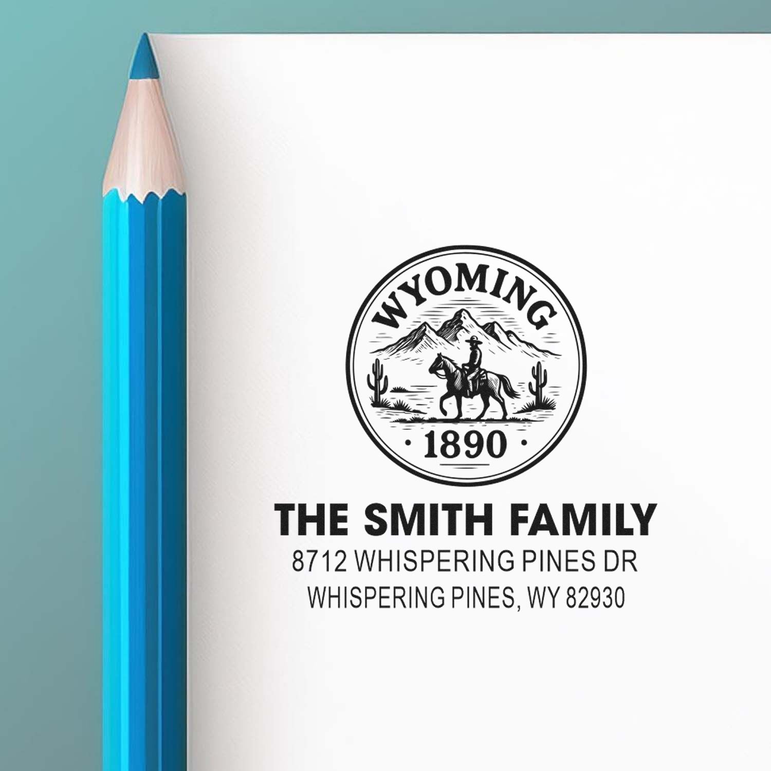 Custom Wyoming family address stamp featuring cowboy, horse, mountains and cacti, displayed beside a blue pencil on white paper.
