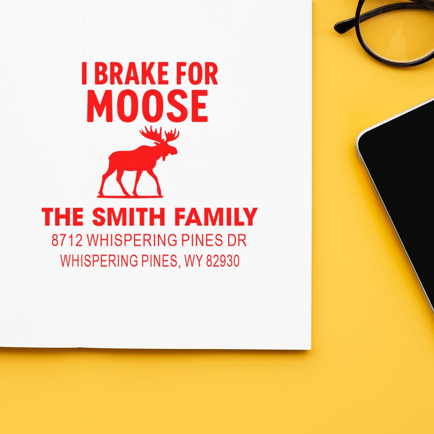 Custom address stamp with red I Brake for Moose moose design and Smith family address on white paper, beside glasses and a smartphone on a yellow desk.