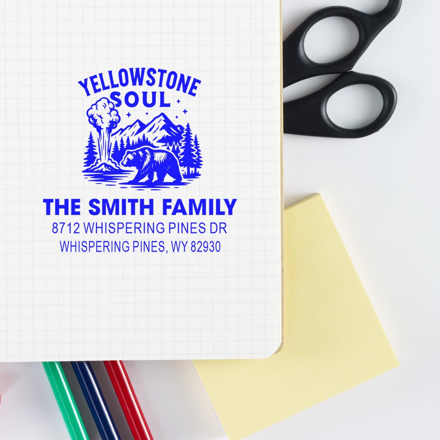 Custom blue Yellowstone Soul family address stamp on notebook with scissors, pens and sticky note, featuring bear, mountains and geyser design.