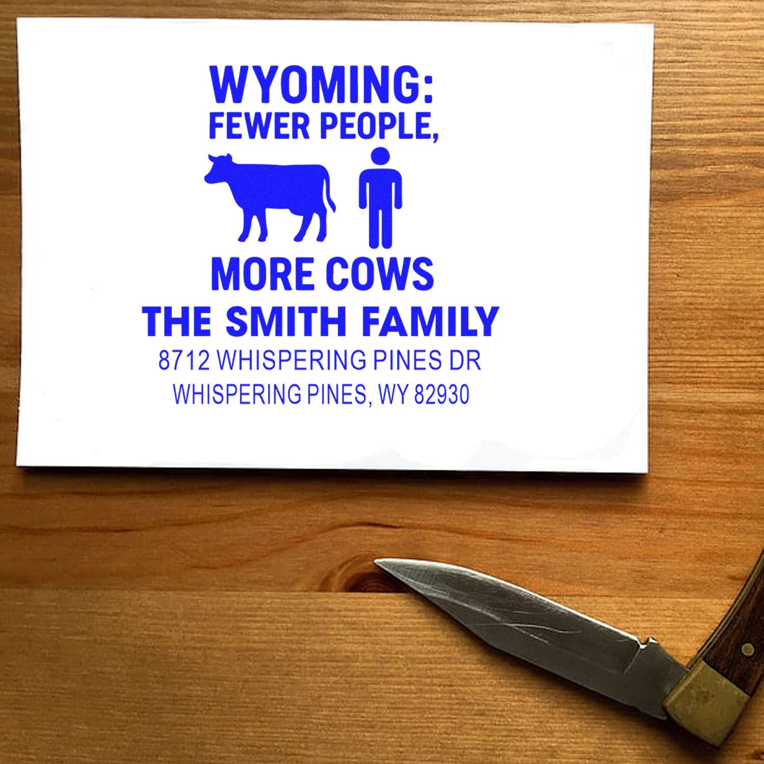 Custom Wyoming address stamp with cow and person graphic, reading Fewer People, More Cows, on white card on a wooden table.