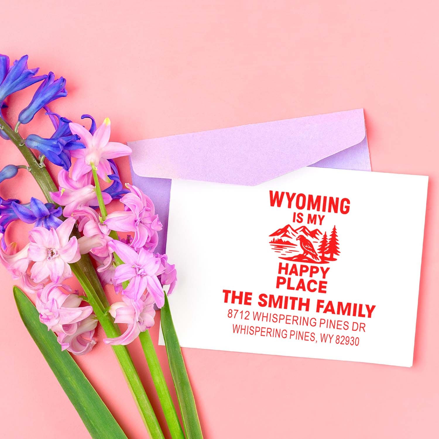 Custom Wyoming return address stamp on a white envelope beside pink and purple flowers on a pastel pink background.