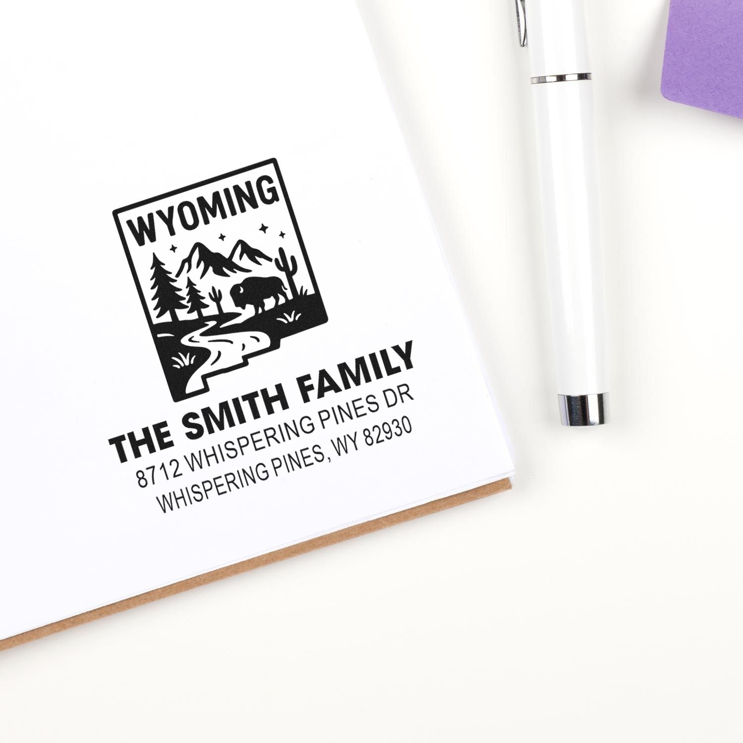 Custom Wyoming family address stamp on white stationery, with mountain, bison and pine design beside a white pen.