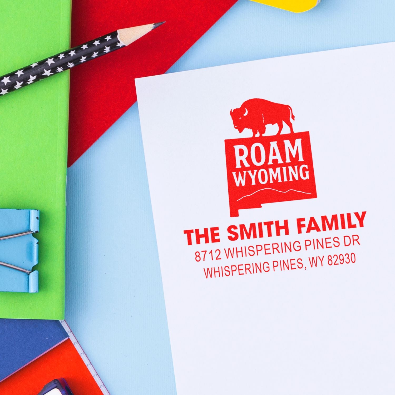 Custom Roam Wyoming address stamp on white card with colorful stationery, star-pattern pencil, and binder clip in a bright flat-lay workspace.