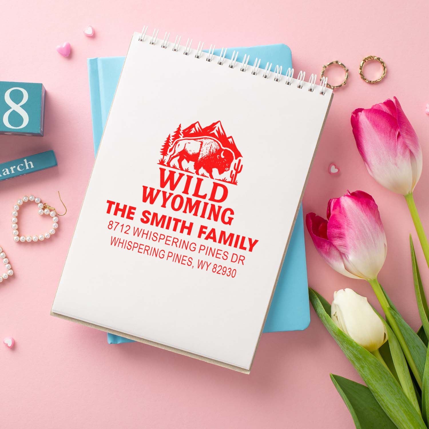 Personalized Wild Wyoming address stamp mockup on a spiral notebook, styled with pink tulips, pastel stationery, and gift decor on a pink background.