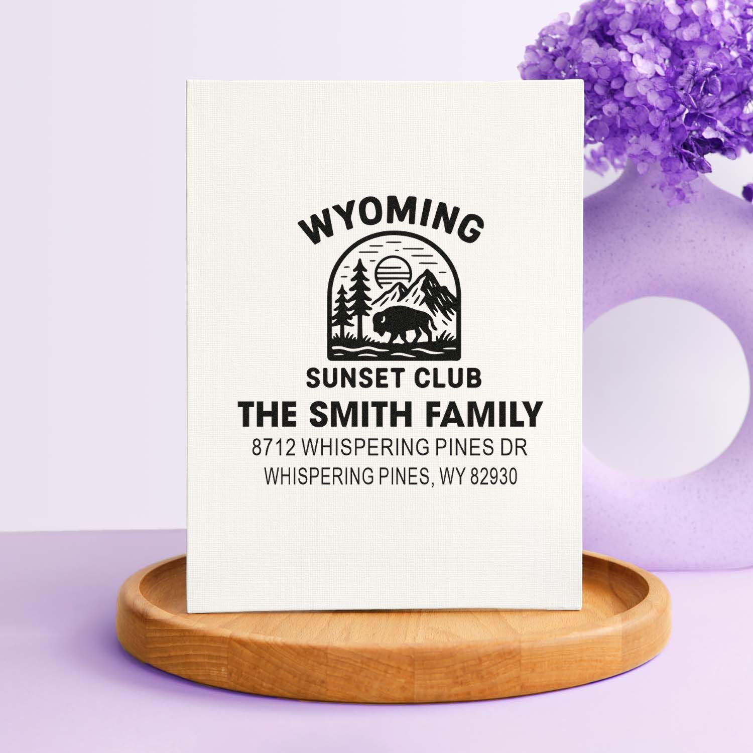 Custom Wyoming Sunset Club address stamp on white canvas, featuring bison, mountains and pine trees, displayed on a wooden stand with purple decor.