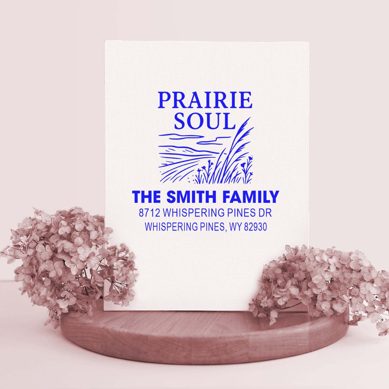 Custom Prairie Soul family address stamp with blue prairie artwork, displayed on a wooden stand beside blush flowers.