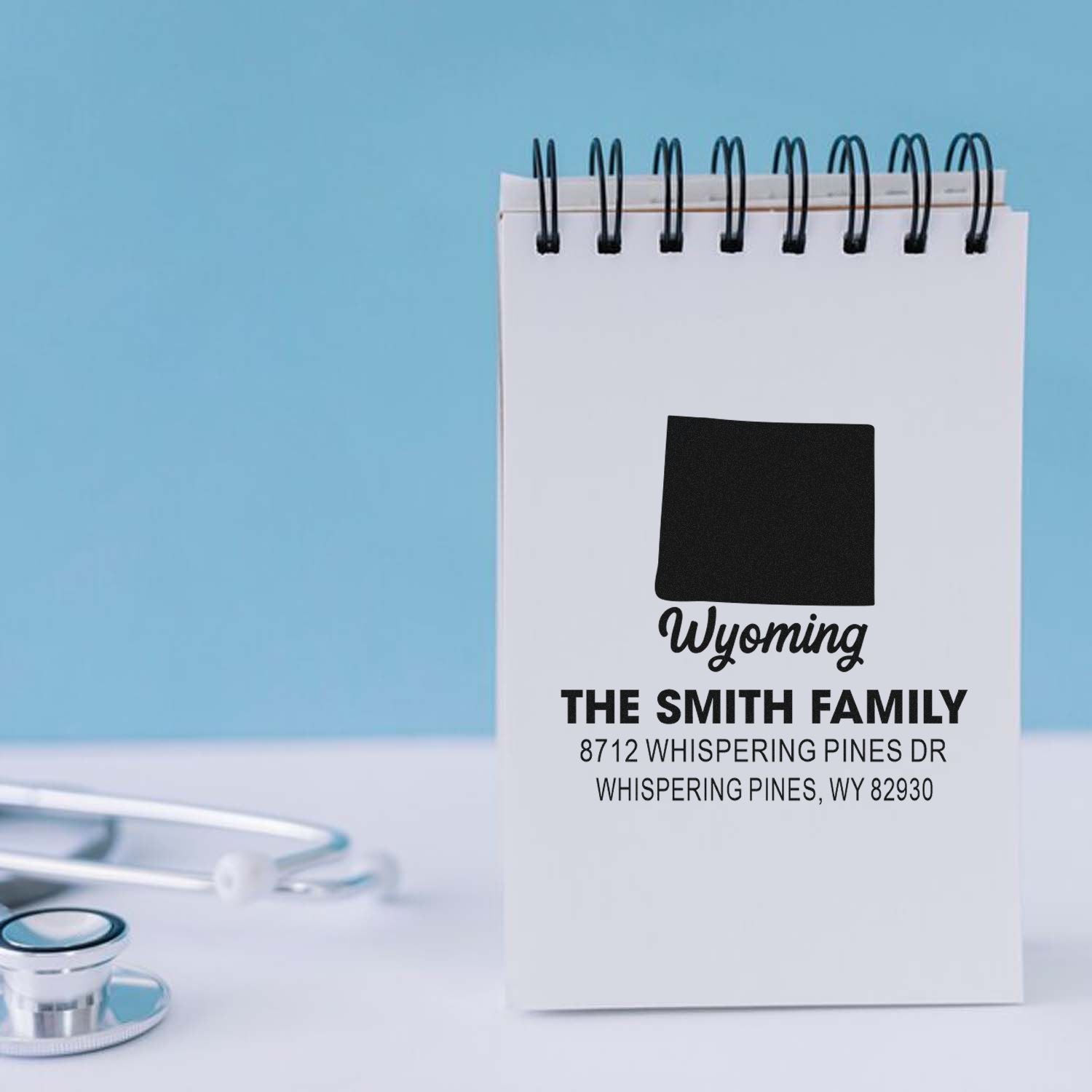 Custom self-inking address stamp on spiral notepad, featuring Wyoming state outline and The Smith Family address on a clean desk beside a stethoscope.