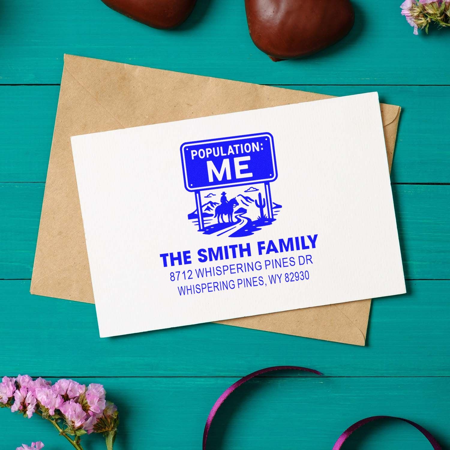 Personalized blue return address stamp reading Population: Me on a white card over a kraft envelope on a turquoise wooden table with flowers.