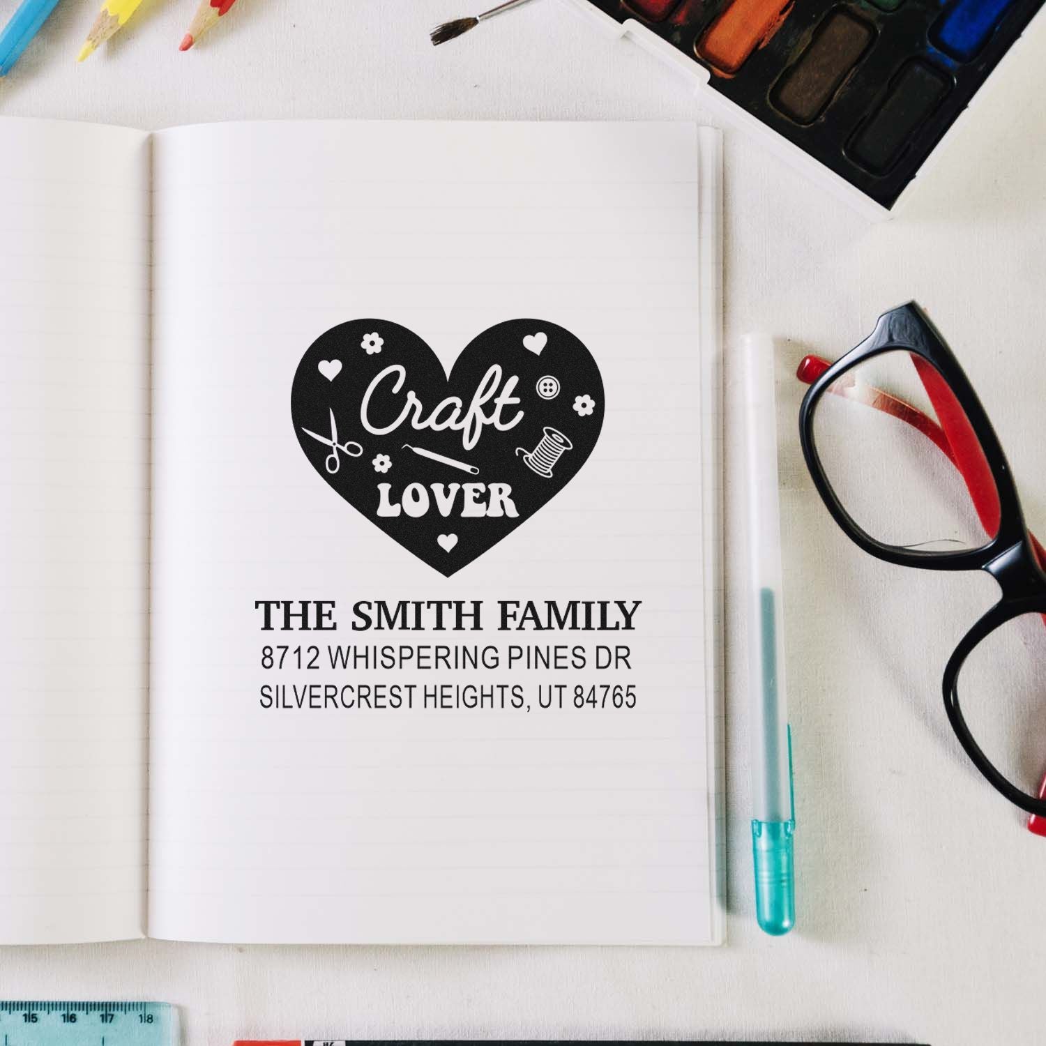 Craft Lover heart stamp with custom family name and address on an open notebook, surrounded by stationery, glasses, and watercolor paints.