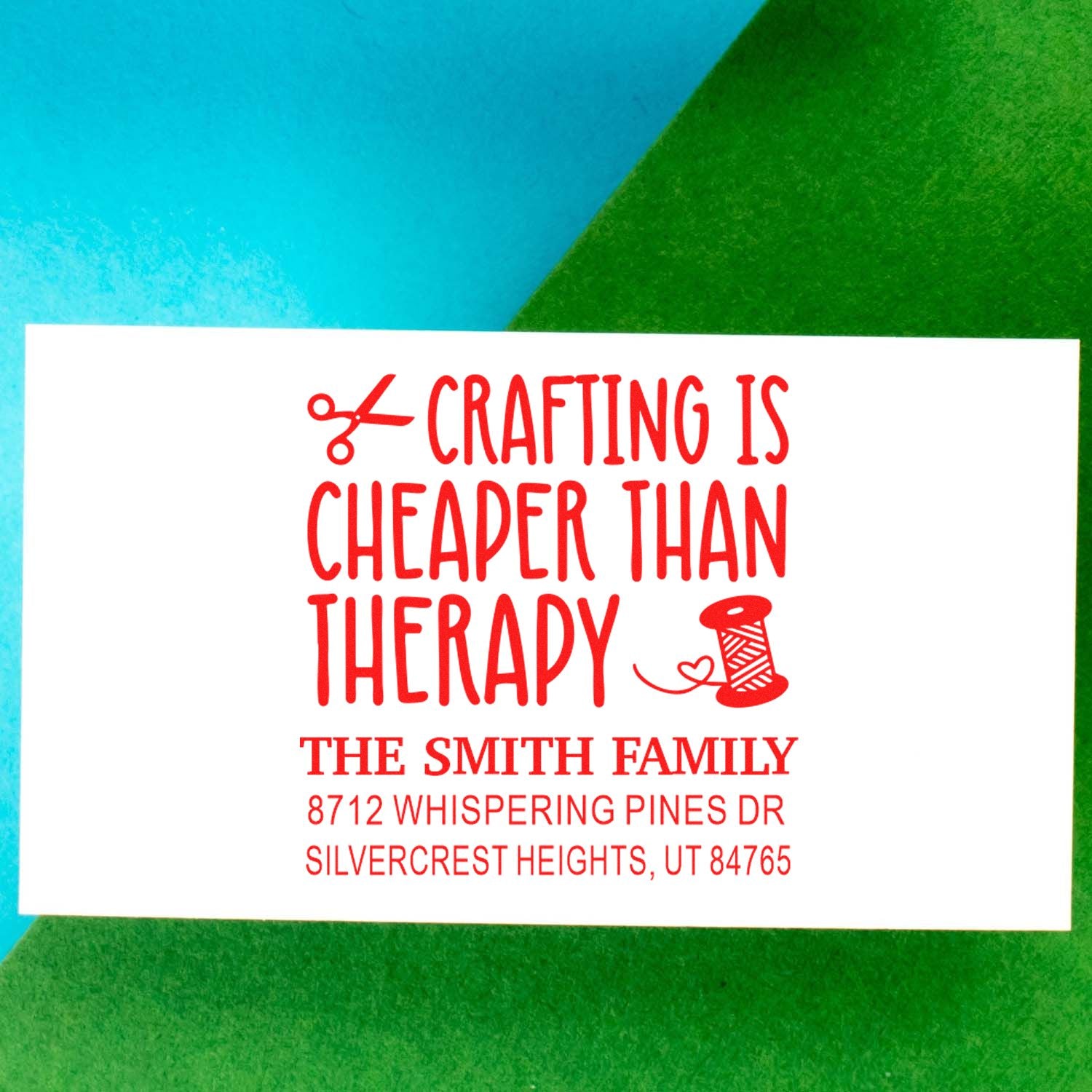 Custom red address stamp reading Crafting Is Cheaper Than Therapy with scissors and thread spool, shown on blue and green background.
