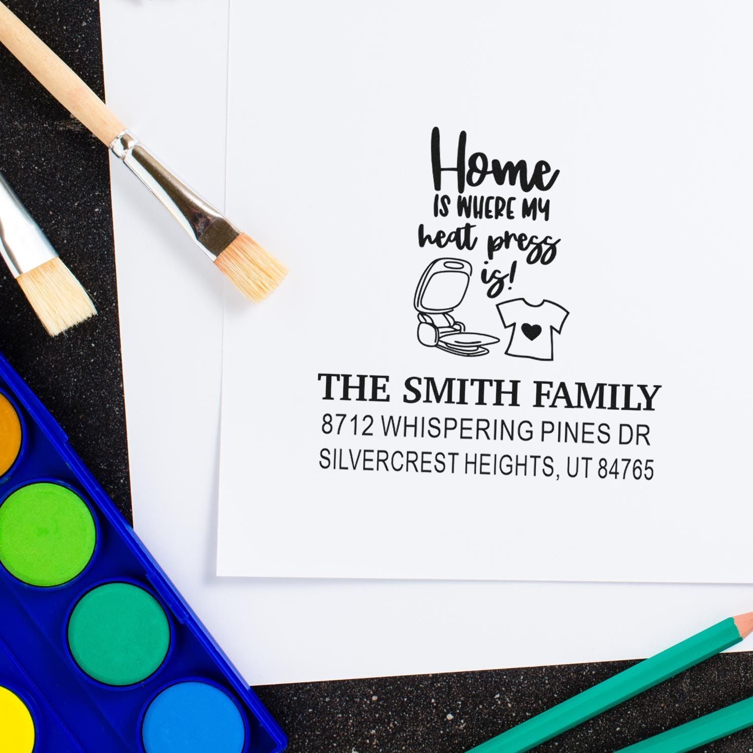 Custom home heat press rubber stamp with family name and address, styled on a craft desk with paintbrushes, watercolor palette, and pencils.