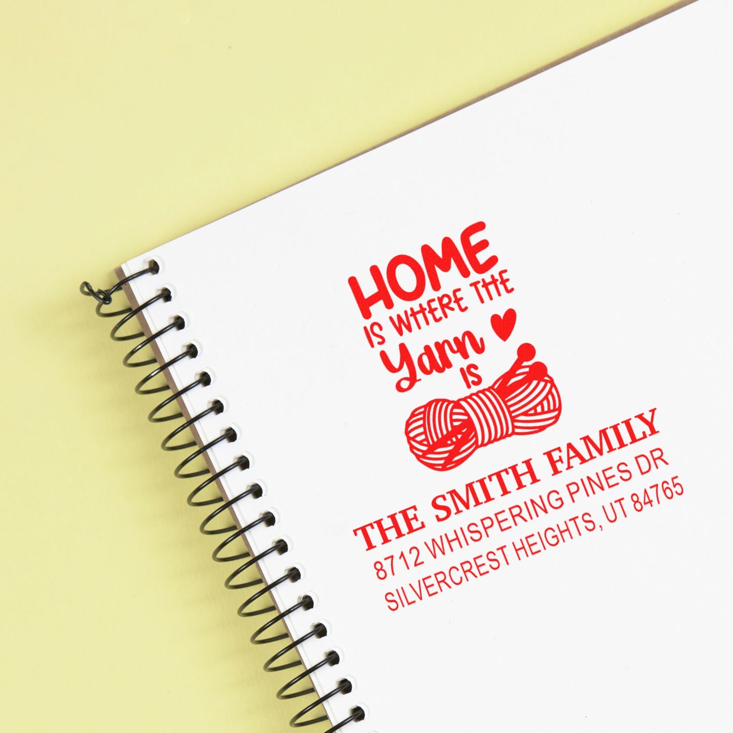 Custom home address stamp on a spiral notebook, featuring Home is where the yarn is with yarn graphic and family name on a white cover against a pale yellow background.