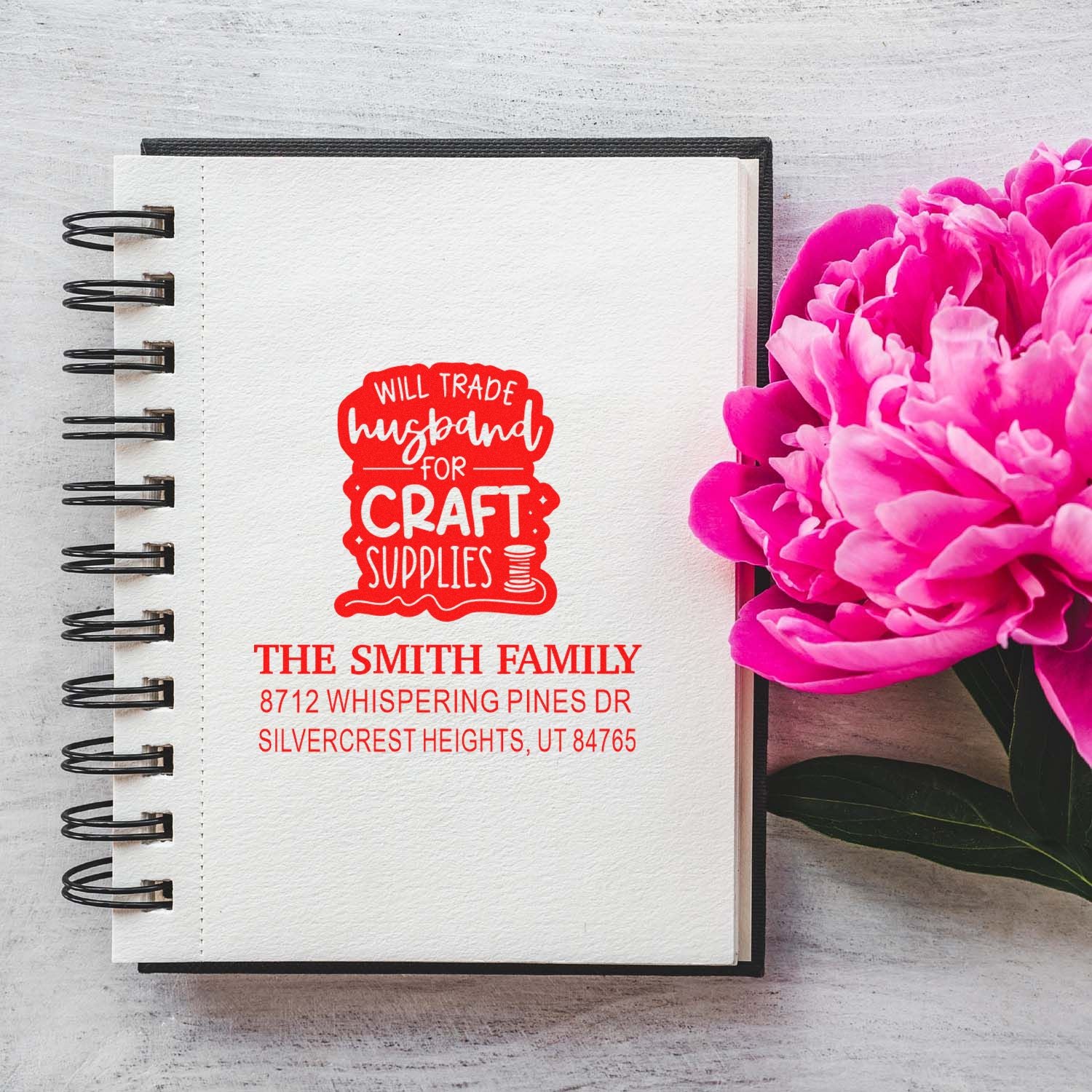 Personalized return address stamp on a spiral notebook beside pink peonies, reading "Will trade husband for craft supplies" with Smith family address.