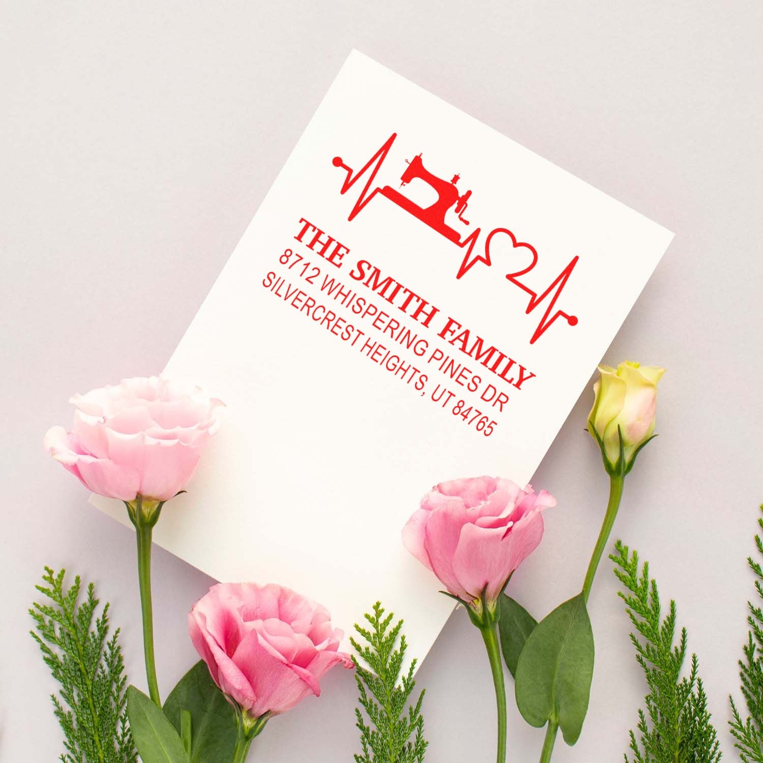 Custom address stamp with sewing machine and heart design, shown on white paper with pink flowers on a soft pastel background.