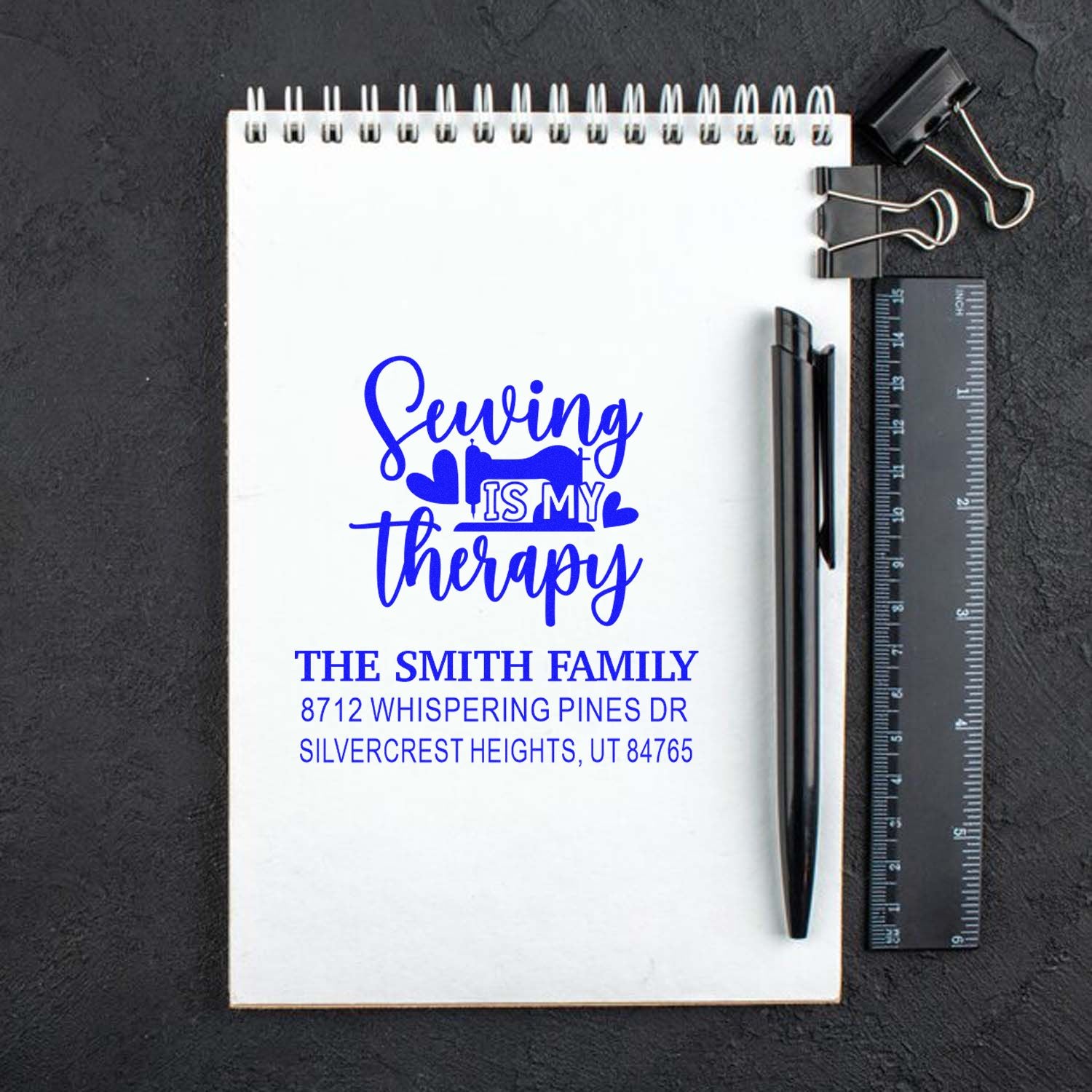 Custom family address stamp with blue Sewing is my therapy design on a notebook, beside a pen, ruler, and binder clips on a dark desk.