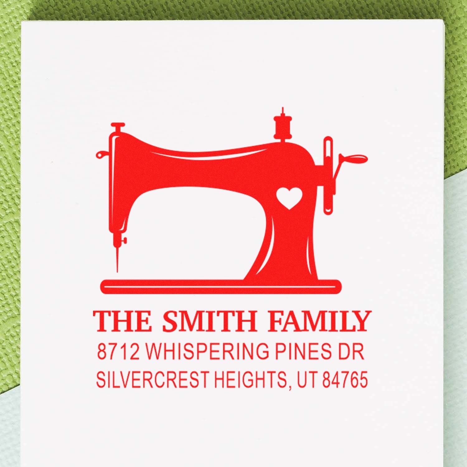Red personalized address stamp with sewing machine and heart design for The Smith Family, on white card against a green textured background.