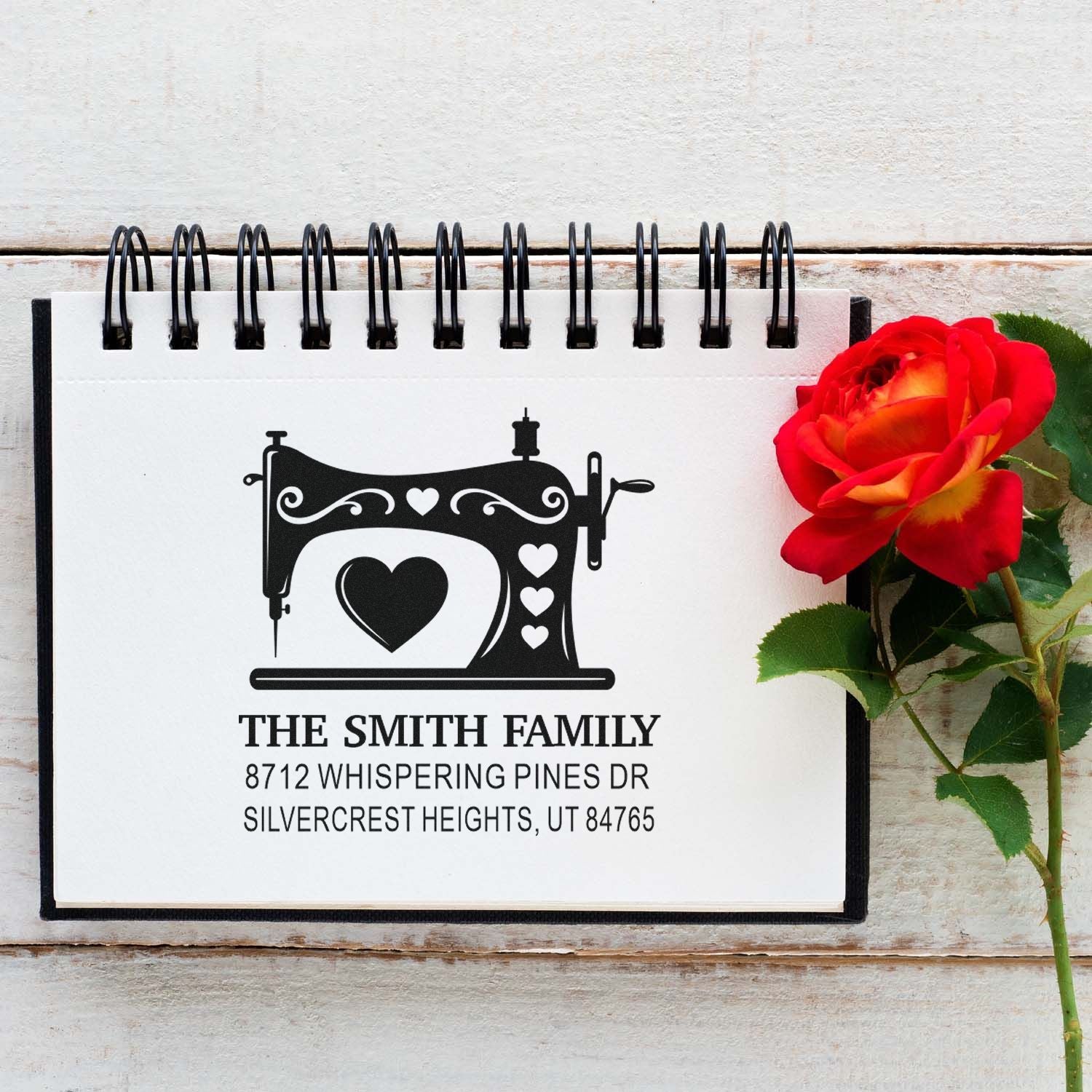 Custom sewing machine return address stamp with heart motif displayed in a spiral notebook beside a red rose on rustic white wood.