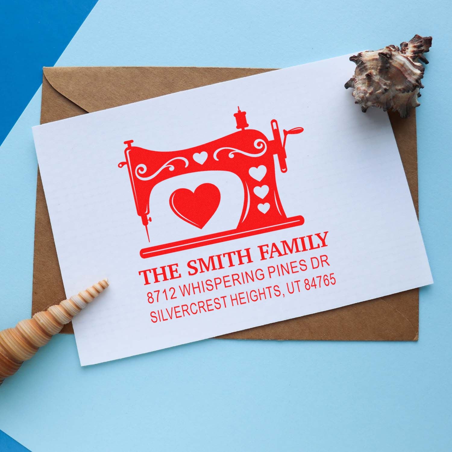 Custom red sewing machine address stamp on white card over kraft envelope, with heart design and family name, beside seashells on a blue background.