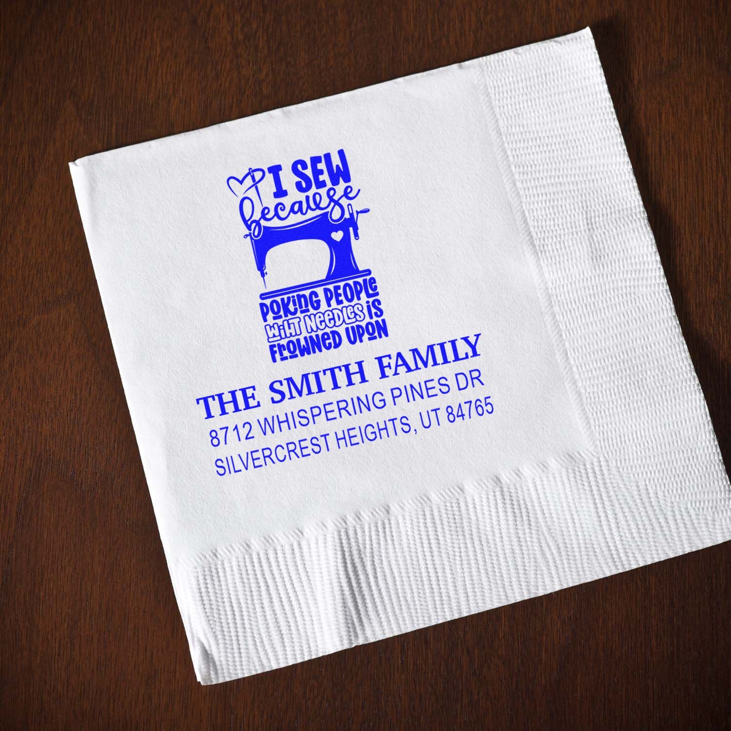 Custom blue sewing machine address stamp on a white napkin, with family name and address, on a dark wood table.