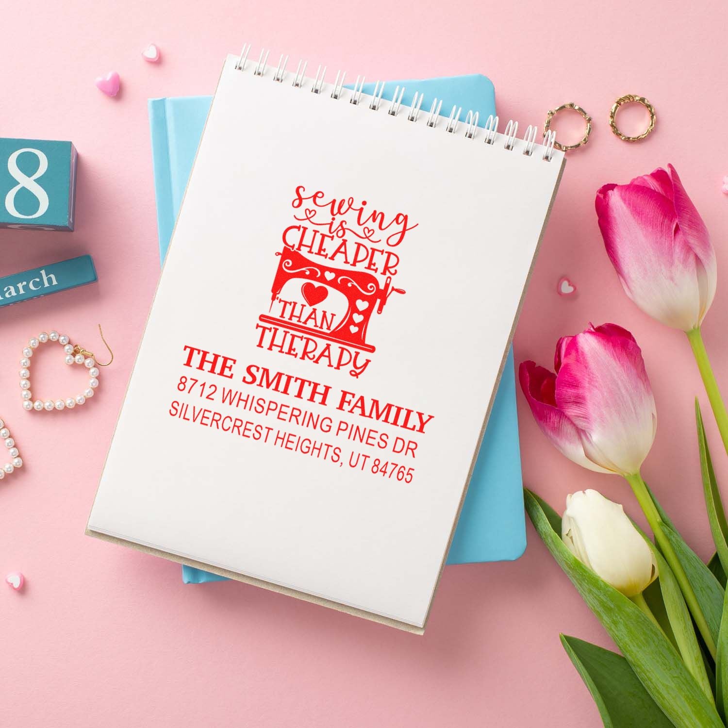 Personalized sewing-themed address stamp on a spiral notebook, styled with pink tulips and pastel stationery on a pink background.