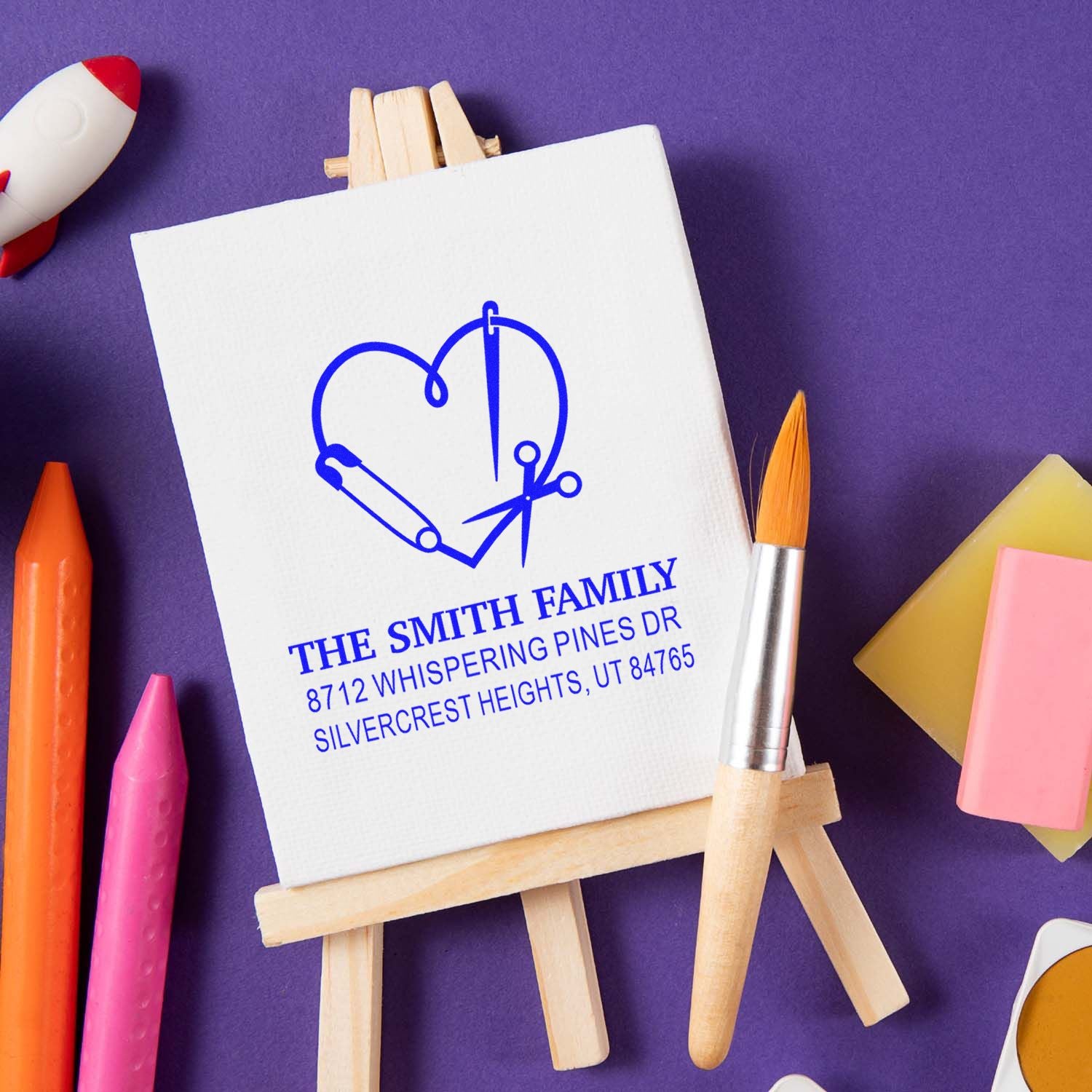 Custom family address stamp on an easel with heart-and-paintbrush design, surrounded by art supplies on a purple background.