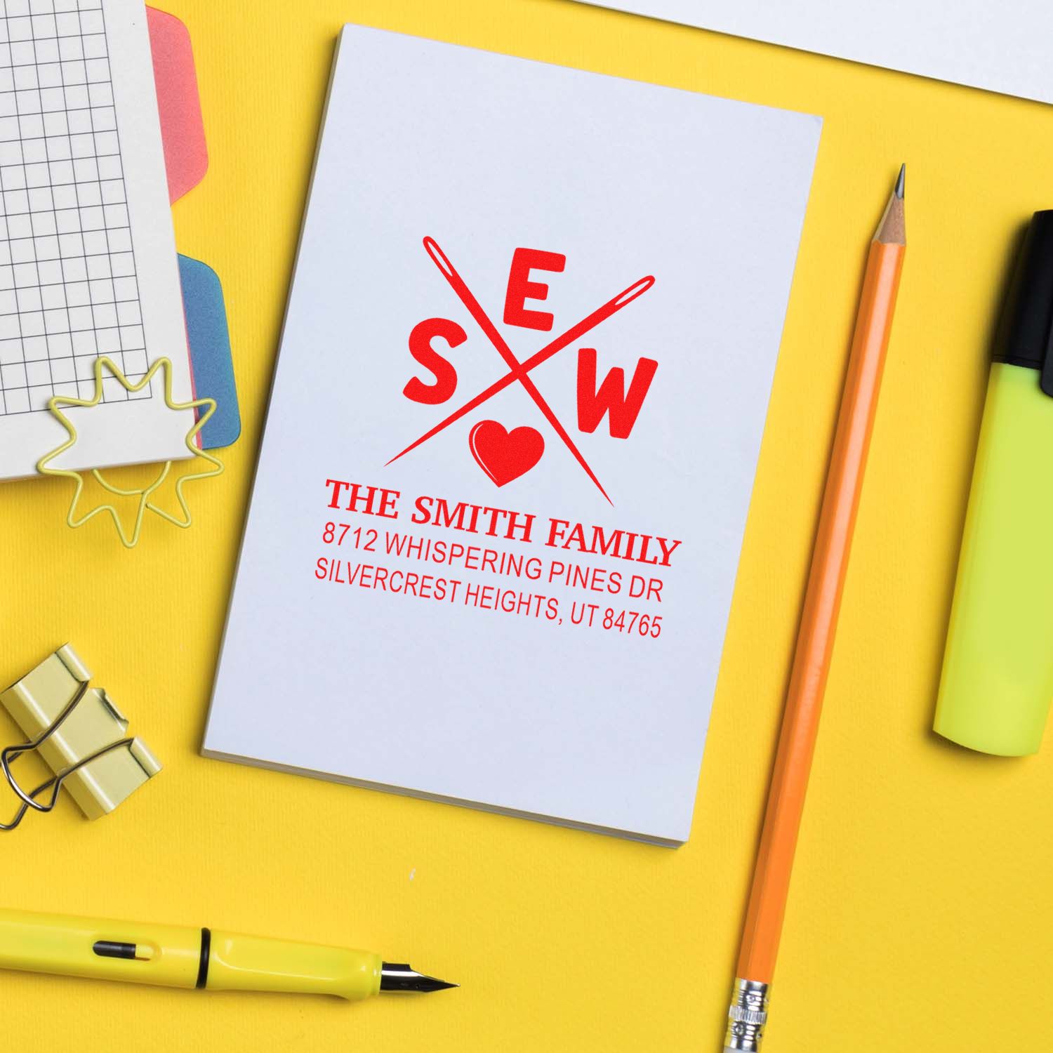 Custom family address stamp with red monogram and address on white card, styled on a yellow desk with pencil, notebook, clips, and stationery.