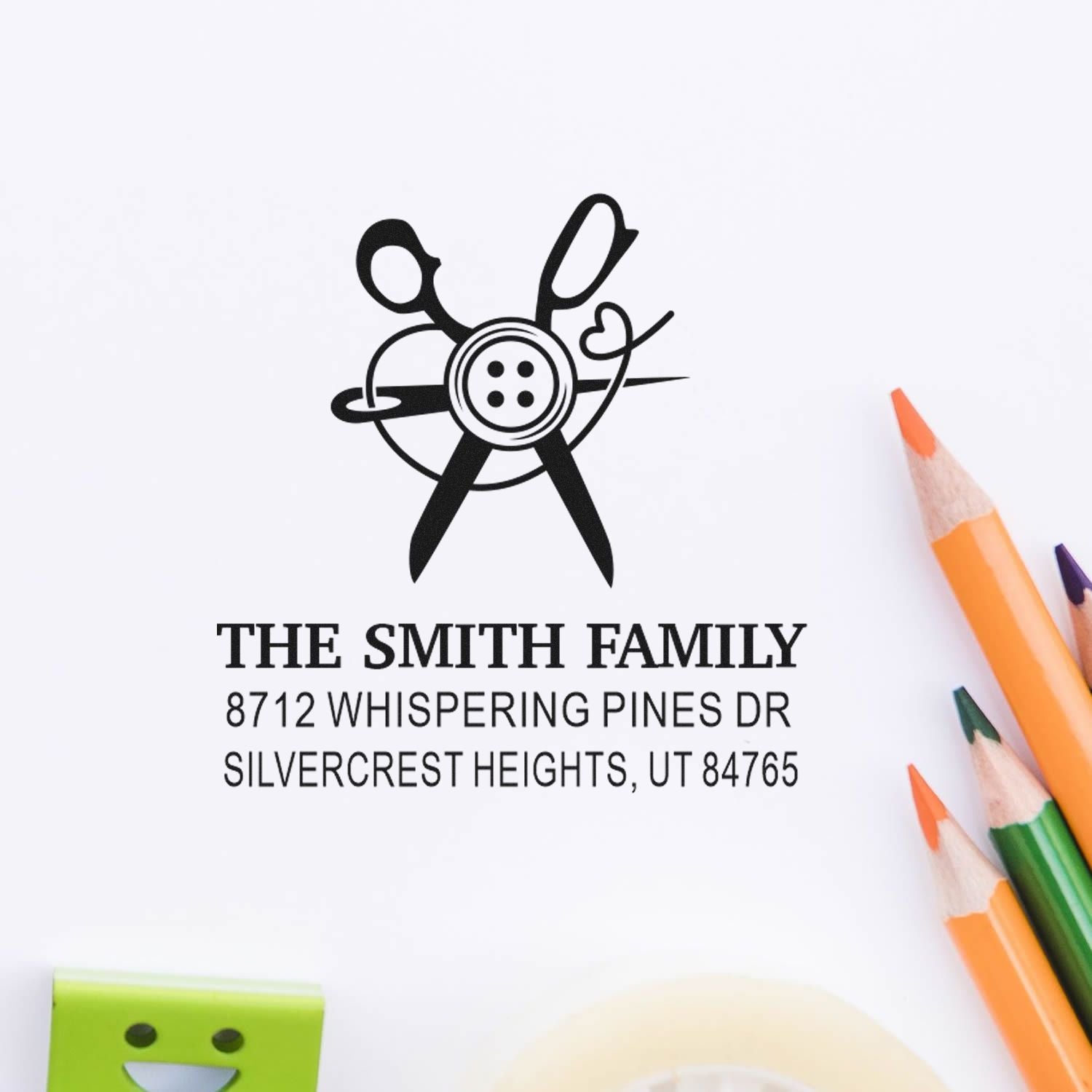 Personalized family address stamp with scissors and button logo, reading The Smith Family, on white paper with colored pencils nearby.