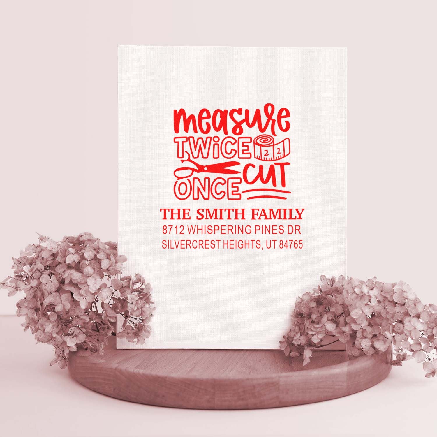 Custom red family address stamp reading measure twice cut once with scissors, displayed on a white backdrop with pink flowers and wooden stand.