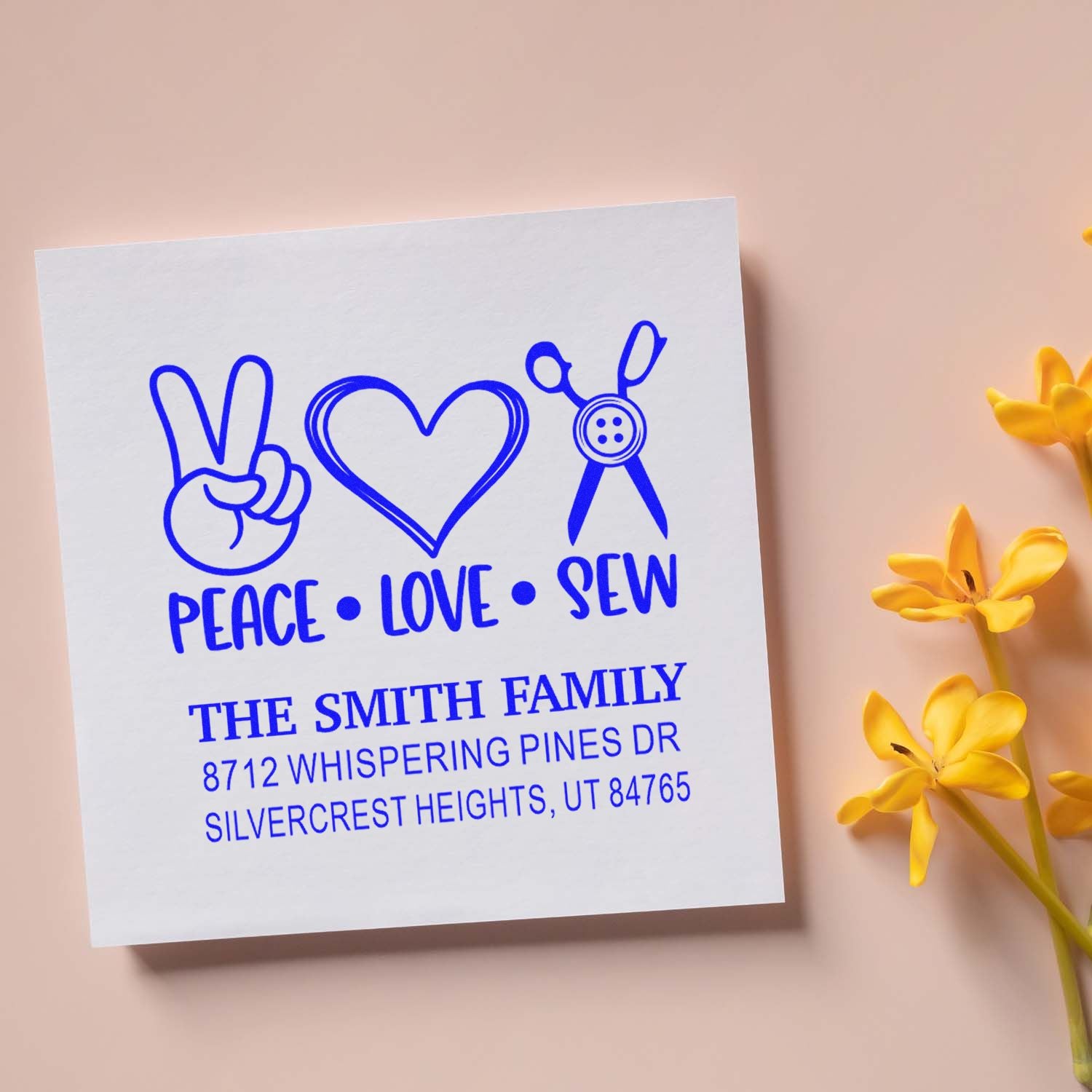 Custom blue Peace Love Sew family address stamp on white card, featuring peace sign, heart, and sewing scissors on a pink background with yellow flowers.
