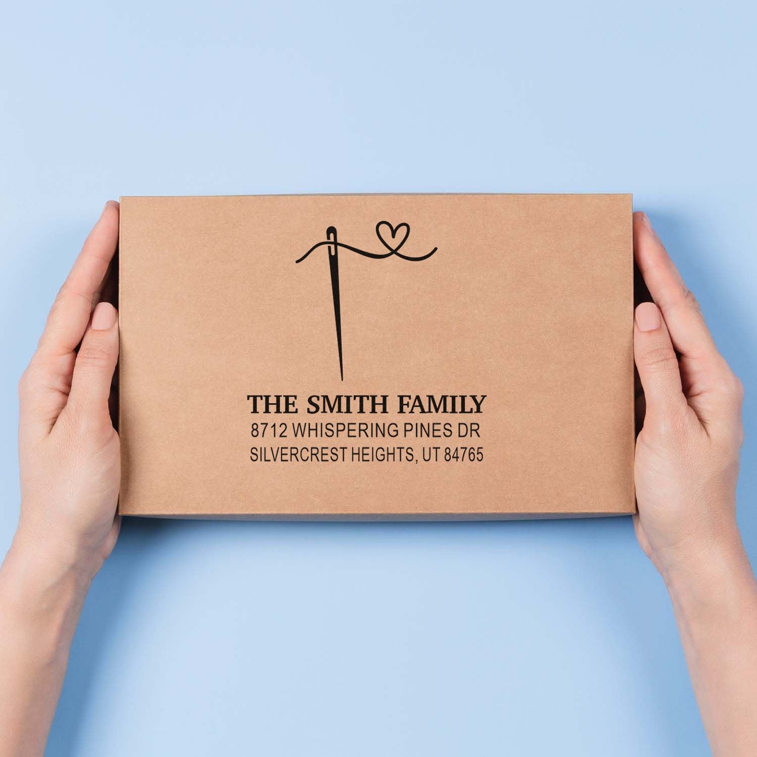 Custom Smith Family return address stamp with heart monogram on kraft mailer box, held in hands against a light blue background.