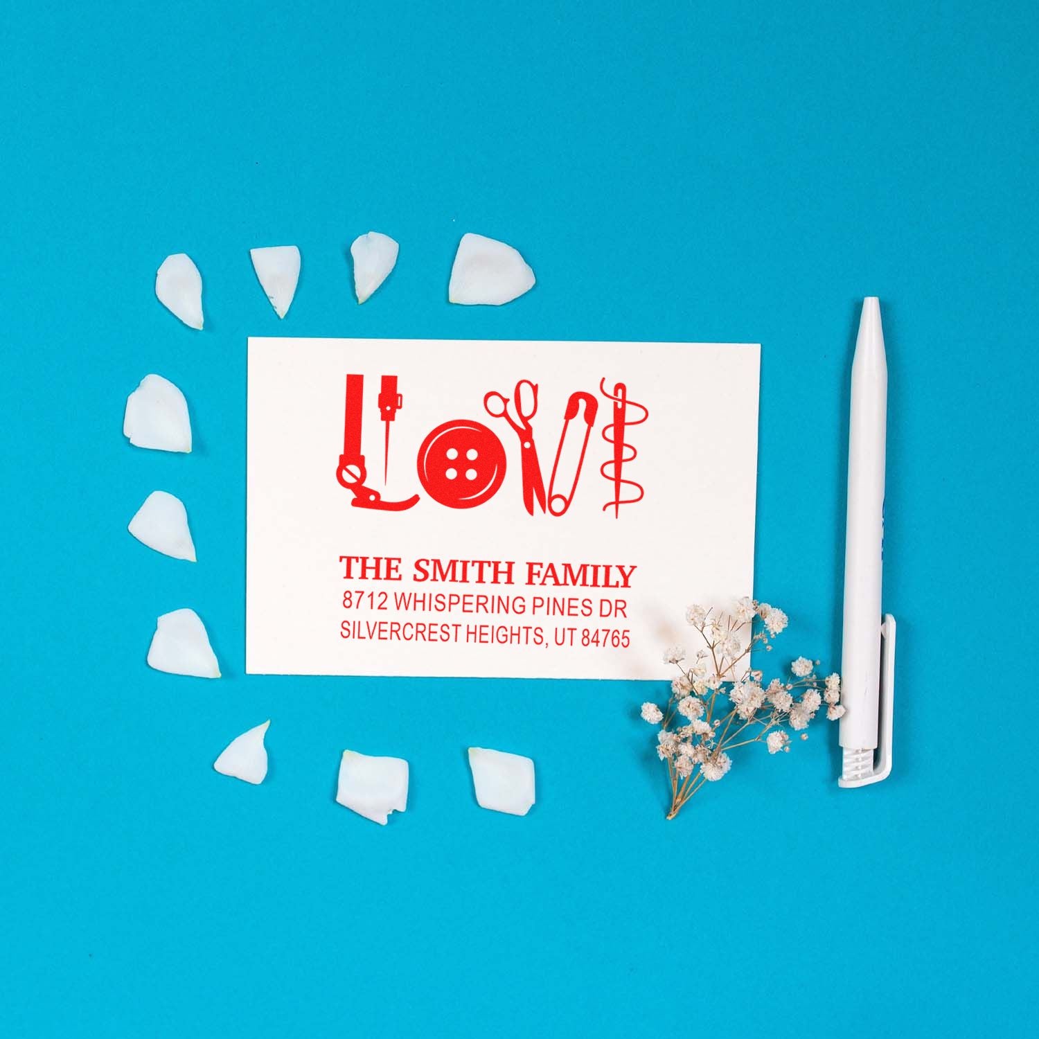 Custom self-inking address stamp featuring red Love made from sewing tools, displayed with a white pen, petals, and dried flowers on a blue background.