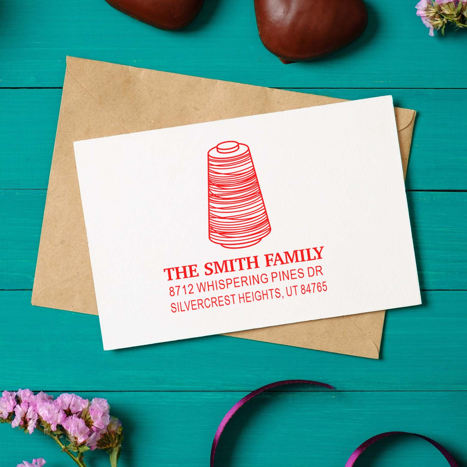 Personalized return address stamp on white card over kraft envelope, with red spool logo and The Smith Family address on a teal tabletop.