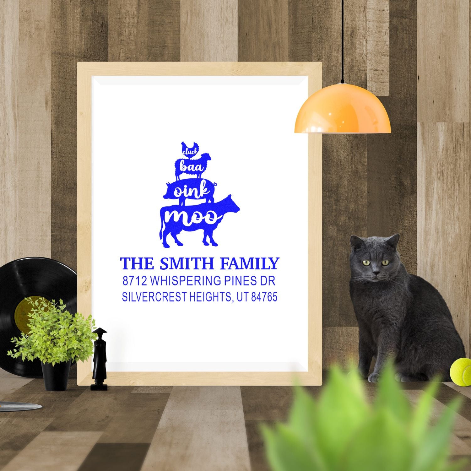 Custom family farm return address stamp mockup in a framed display, with duck, sheep, pig and cow artwork on a wood-paneled tabletop.