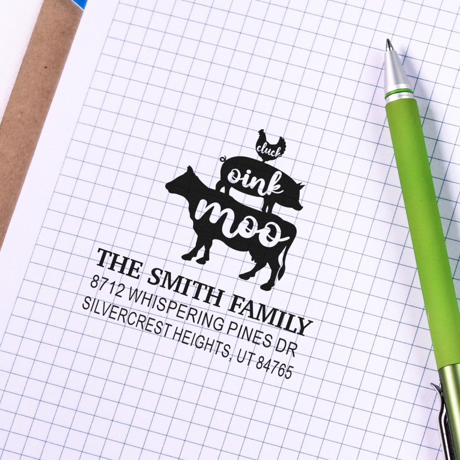 Custom address stamp impression with cow and chicken design on graph paper, beside a green pen, featuring The Smith Family address.