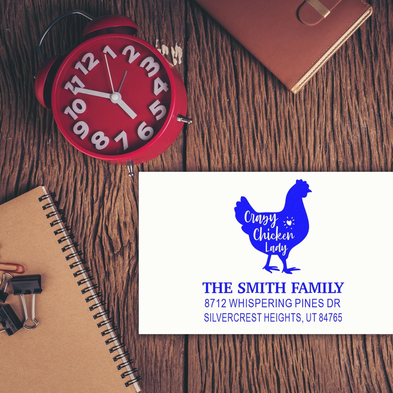 Personalized return address stamp with blue chicken silhouette for The Smith Family on a rustic wood desk beside a red alarm clock and stationery.