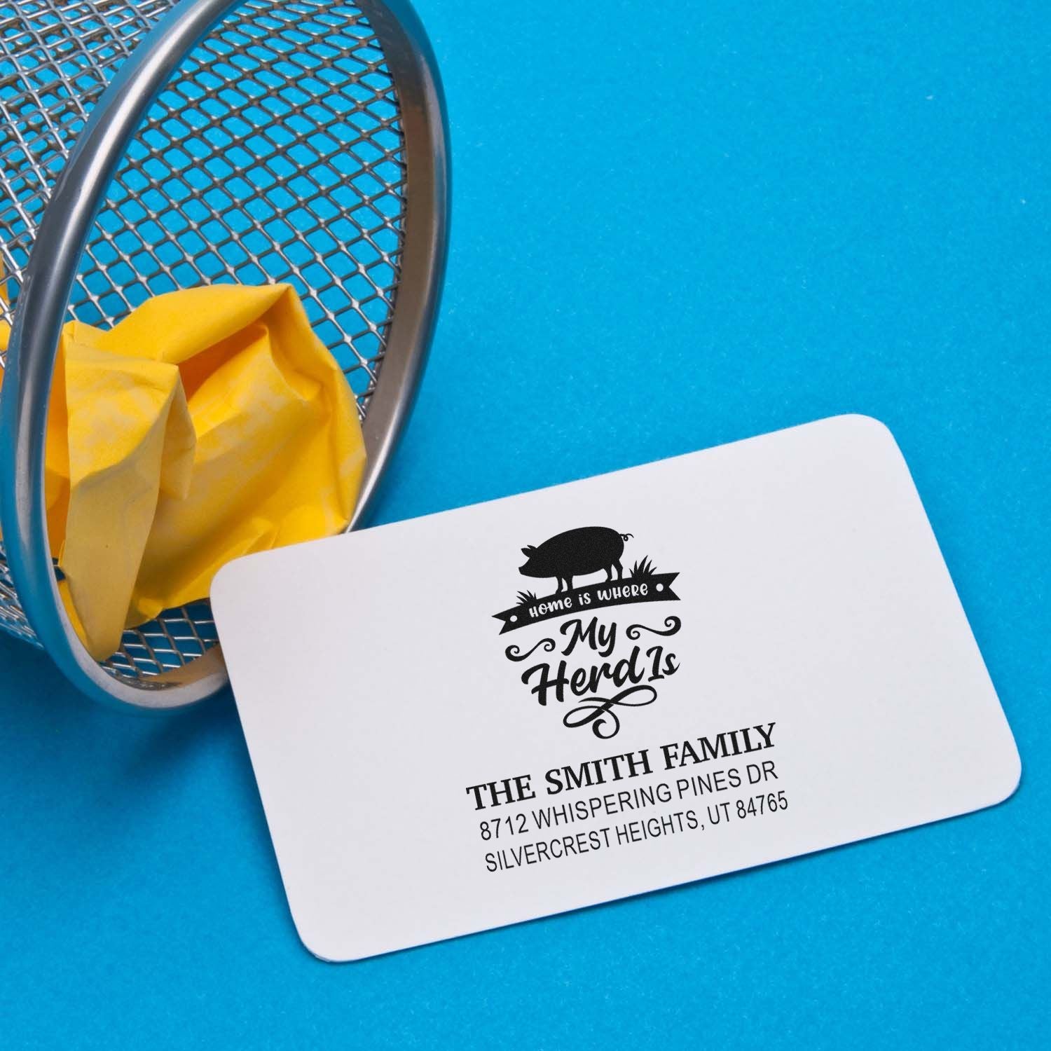 Custom address stamp with pig silhouette and Home Is Where My Herd Is design, beside a mesh tray on a blue background.